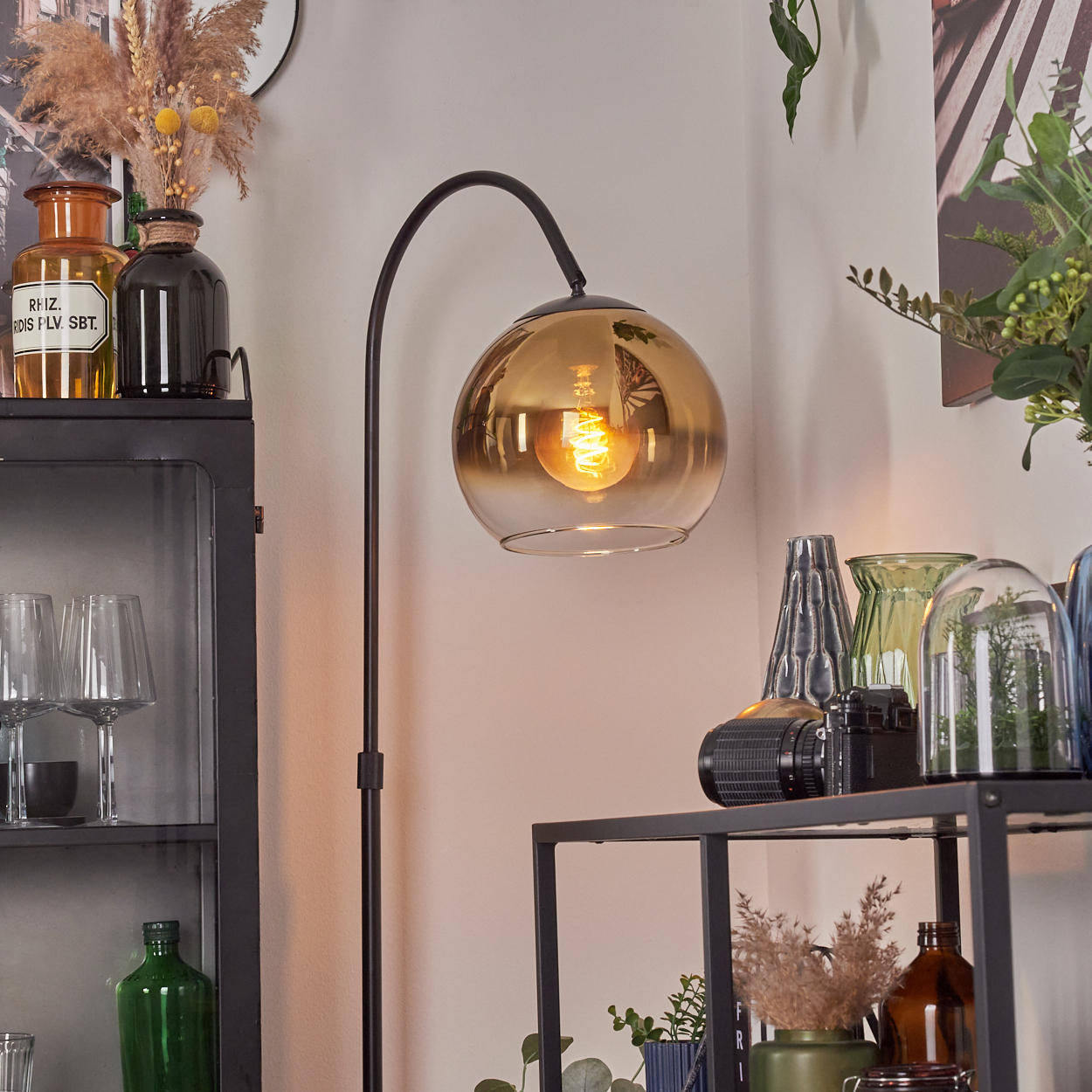 Ein modernes Zimmer mit einer schwarzen Stehlampe mit goldfarbenem Glasschirm. Regale mit Vasen und Dekoration im Hintergrund.