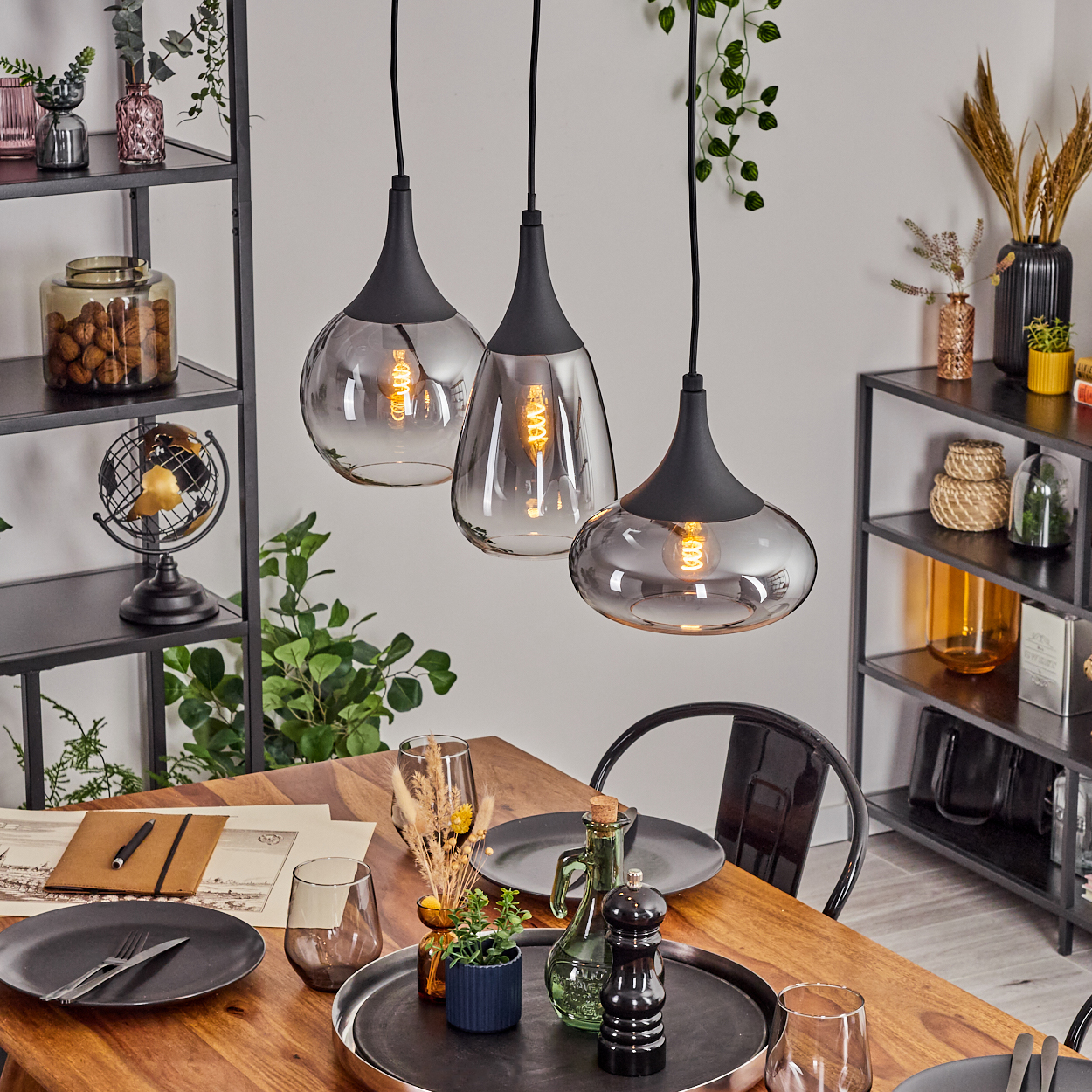 Esszimmer mit Holztisch, schwarzen Pendelleuchten und Regalen. Tisch mit Tellern und Gläsern gedeckt. Pflanzen und Dekorationen vorhanden.