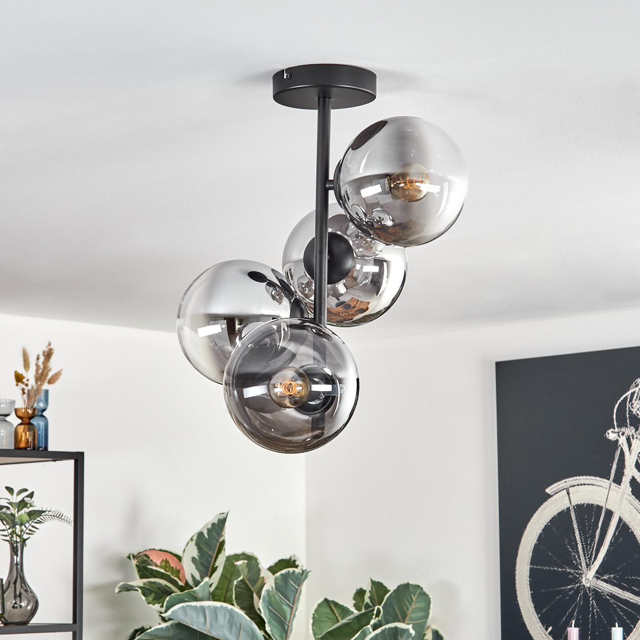 Eine moderne Deckenleuchte mit vier Glaskugelleuchten hängt von einer weißen Decke. Pflanzen und ein Fahrradkunstwerk sind im Hintergrund.