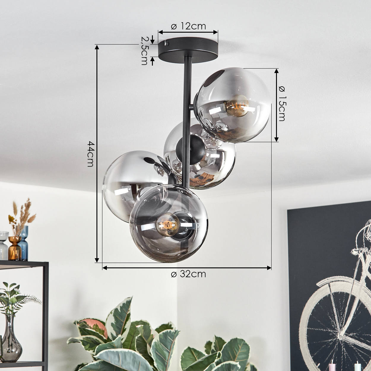 Eine moderne Deckenlampe mit vier Glaskugeln und schwarzen Armaturen. Abmessungen sind angegeben. Im Hintergrund sind Pflanzen und ein Fahrradkunstwerk zu sehen.