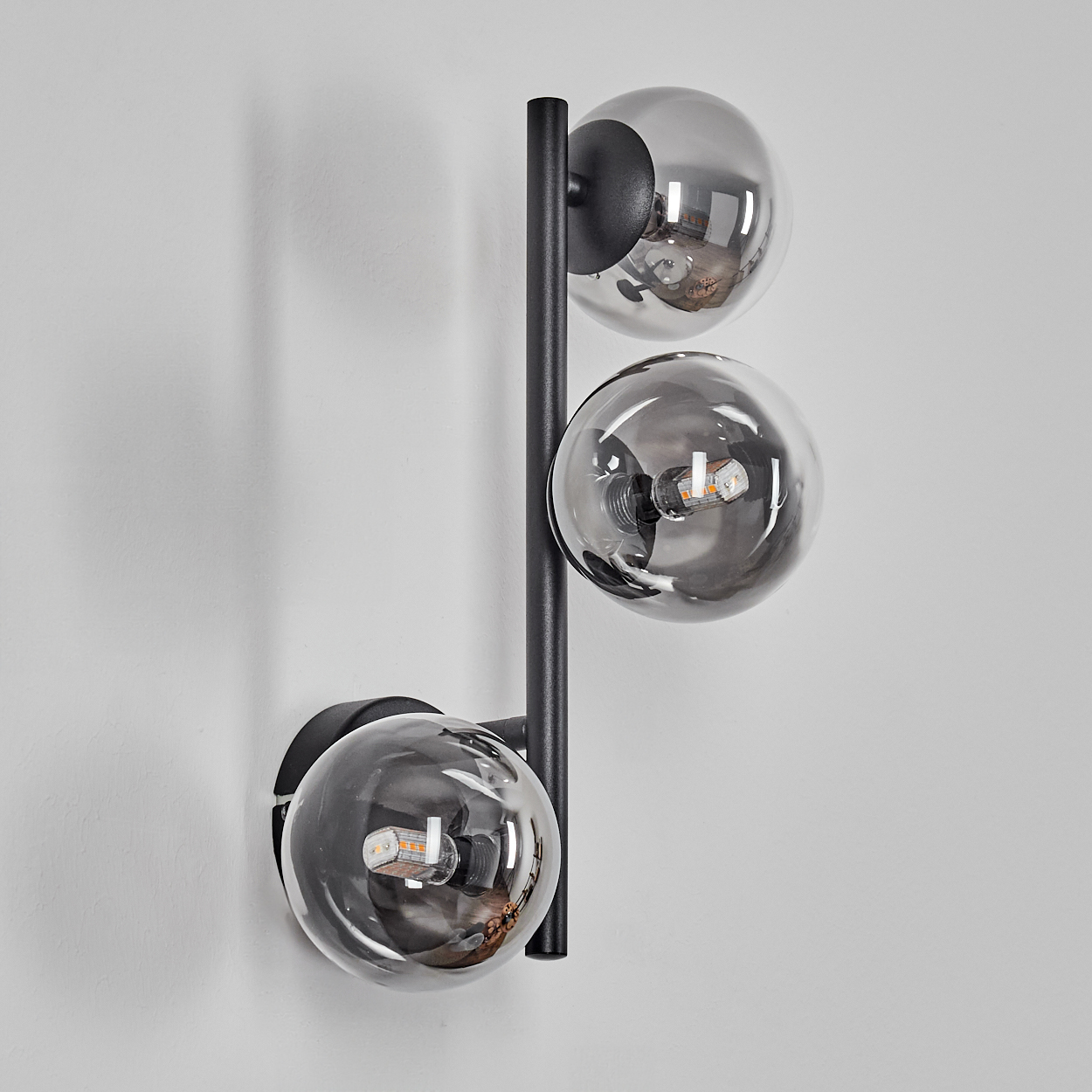 Eine moderne Wandleuchte mit drei kugelförmigen Schirmen. Die Schirme sind aus grauem Glas. Die Lampe ist auf einem schwarzen Metallrahmen montiert.