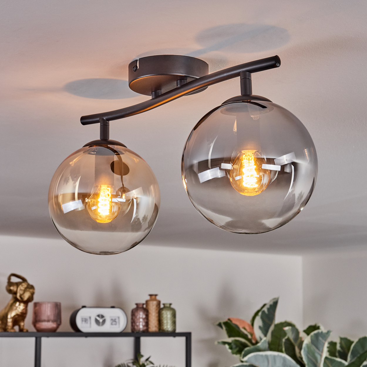 Eine moderne Deckenleuchte mit zwei kugelförmigen Schirmen. Jede Lampe hat eine brennende Glühbirne. Im Hintergrund sind Regale mit Dekoration zu sehen.