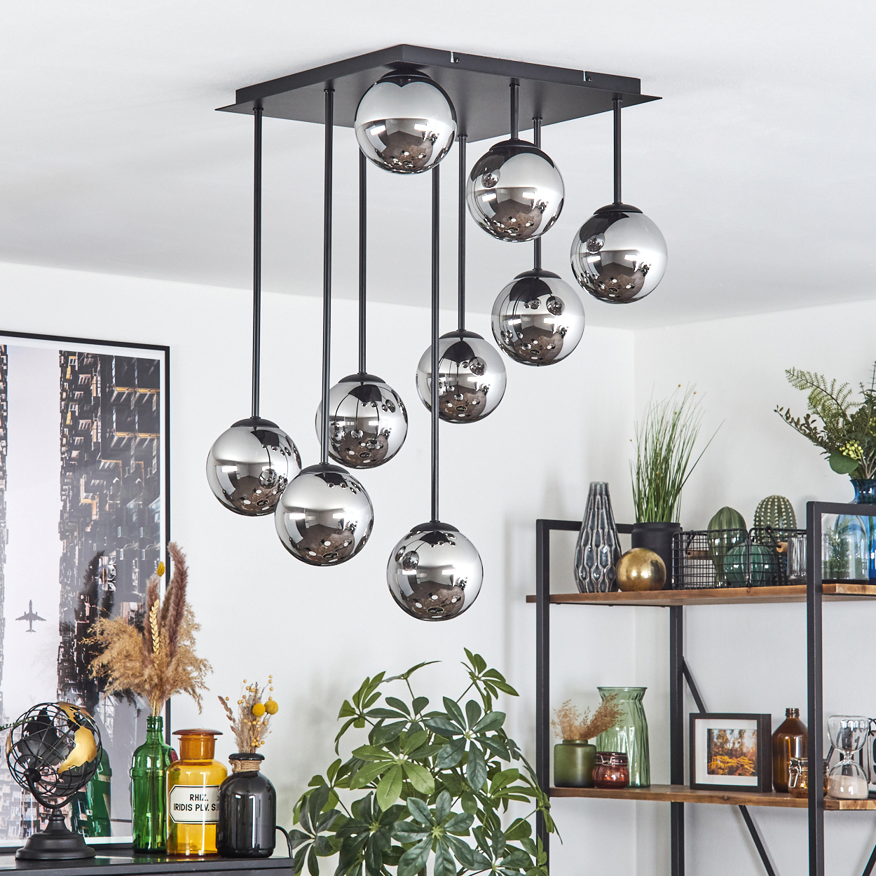Eine moderne Deckenleuchte mit kugelförmigen Chromlampen hängt von der Decke. Regale zeigen Pflanzen und Dekoration. Ein gerahmtes Stadtbild ist an der Wand.