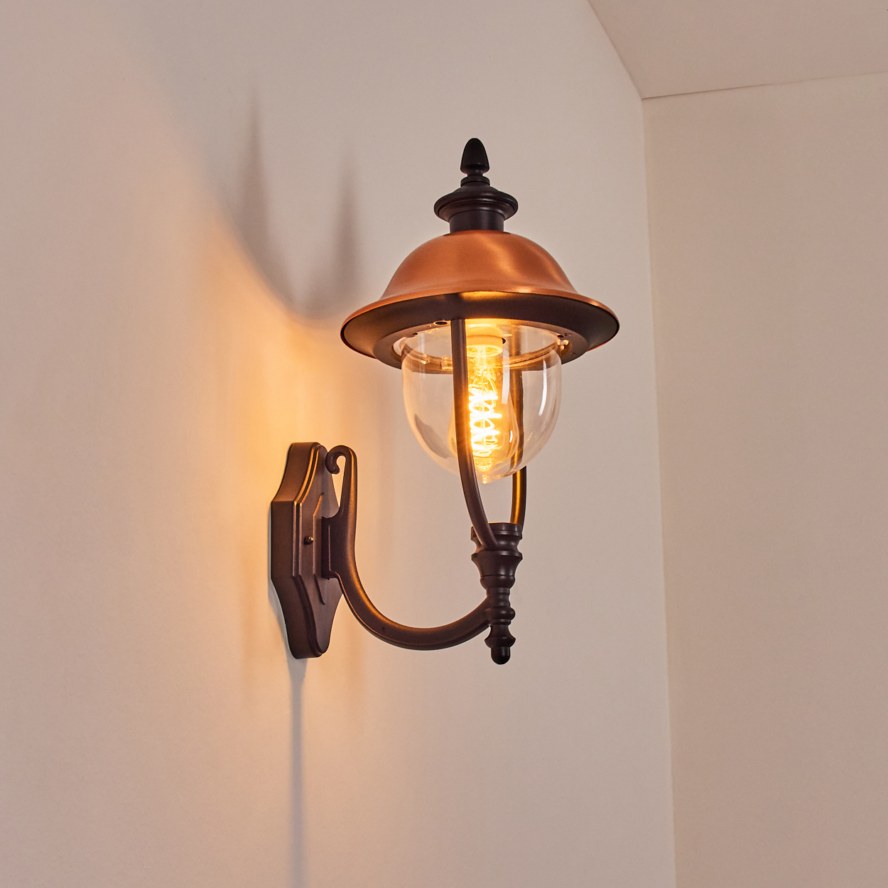 Eine dekorative Wandlampe mit Kupferschirm und klarem Glas. Sie hat einen warmen Schein an einer weißen Wand.