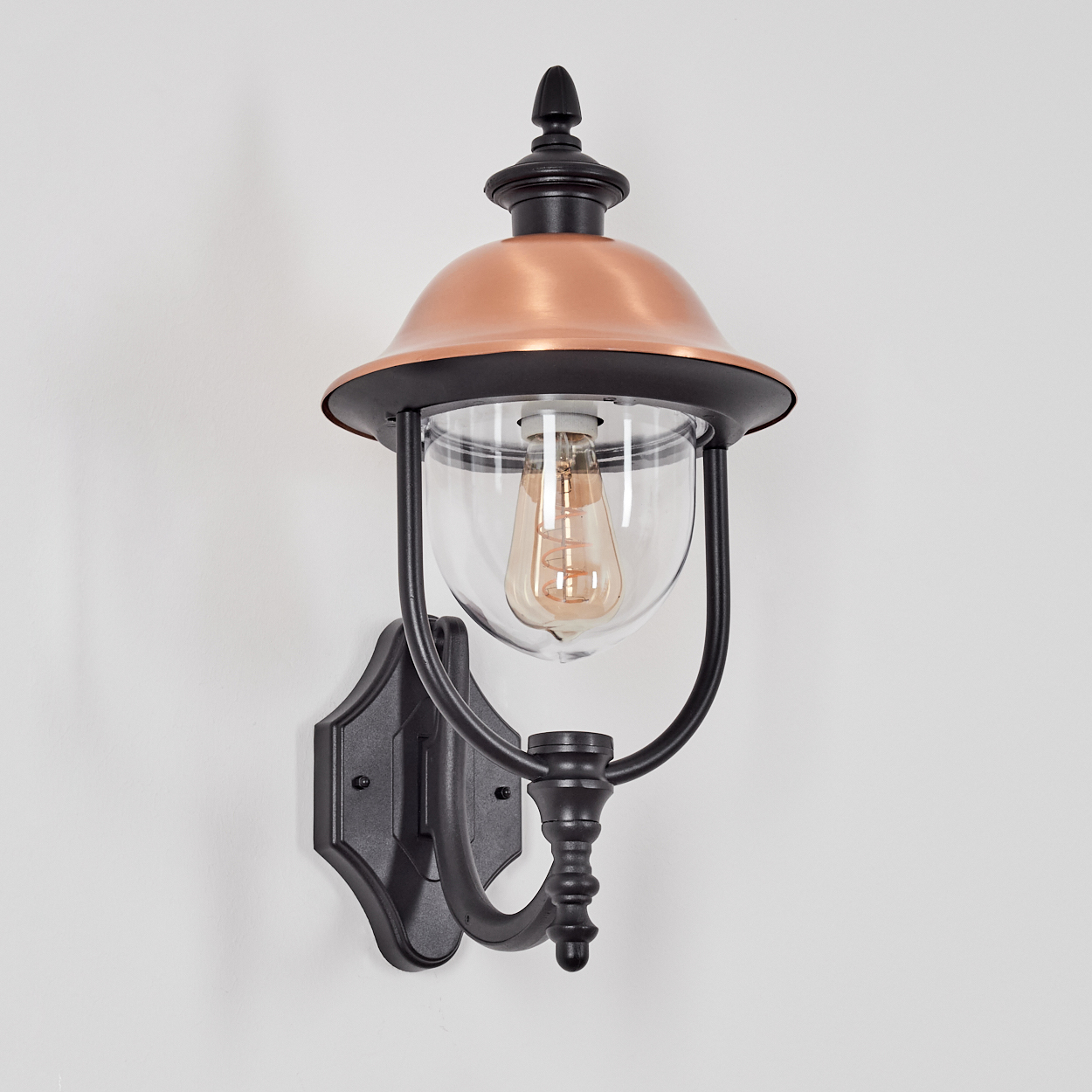 Eine an der Wand montierte Außenlampe mit kupferfarbenem Oberteil, schwarzem Rahmen und einer Glaskuppel mit Glühbirne.