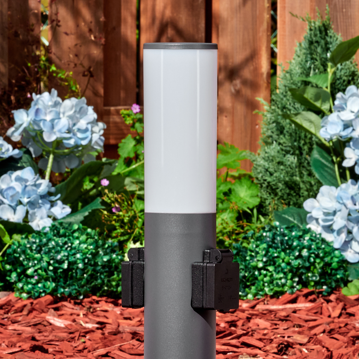 Eine graue Außenlampe mit weißem Oberteil steht in einem Garten. Blaue Blumen, grüne Büsche und ein Holzzaun sind im Hintergrund.