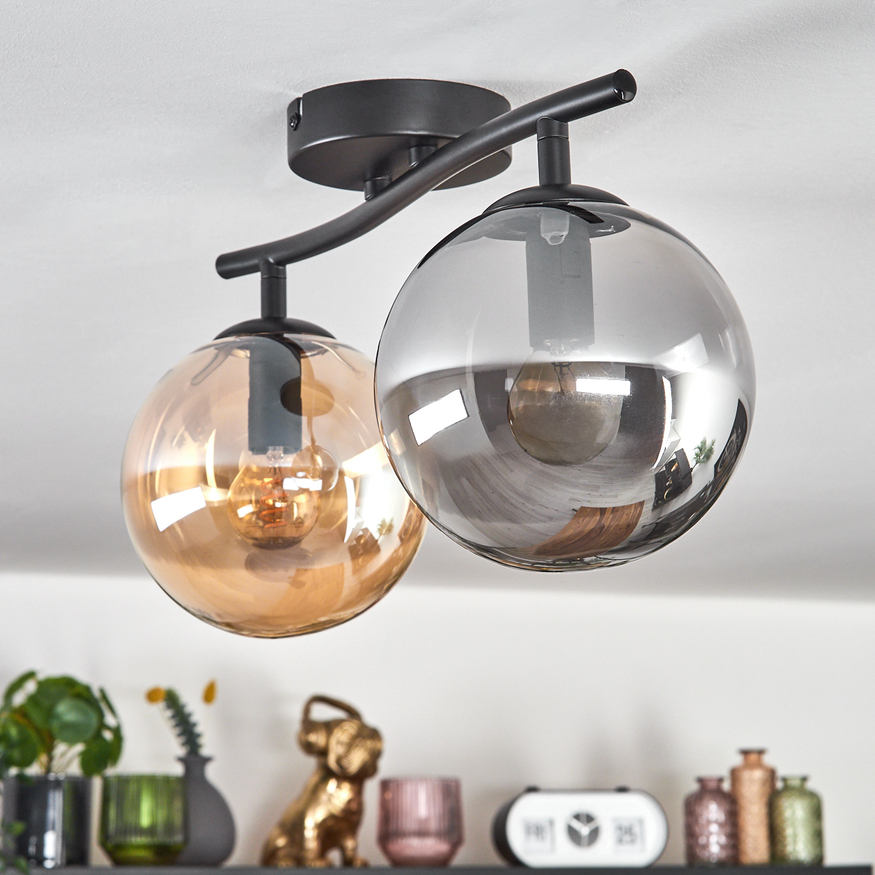 Moderne Deckenlampe mit zwei kugelförmigen Schirmen, einer bernsteinfarben, einer grau. Darunter ein Regal mit Deko, darunter goldene Bulldogge.