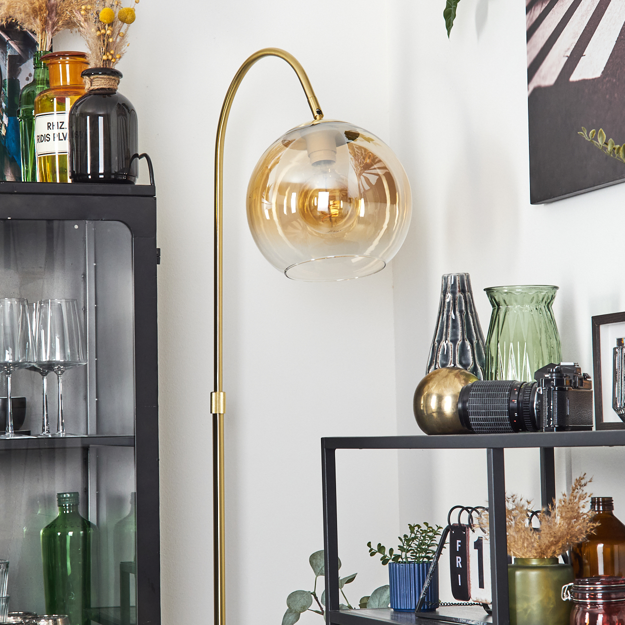 Eine goldene Lampe mit einer bernsteinfarbenen Kugel. Regale beherbergen Vasen, eine Kamera und andere Dekorationsgegenstände. Links ein schwarzer Schrank mit Glastüren.