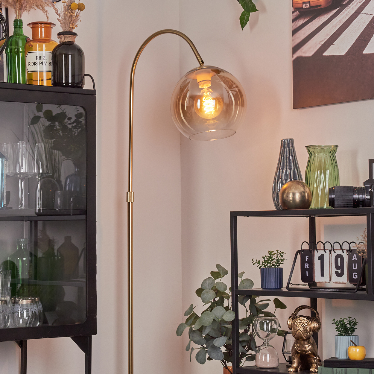Eine goldene Stehlampe mit kugelförmigem Schirm beleuchtet eine Ecke. Regale zeigen Vasen, Pflanzen und einen Kalender. Eine Bulldoggenstatue steht daneben.