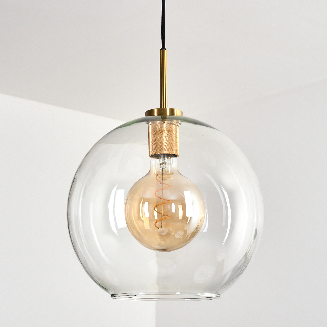 Eine durchsichtige Glaskugel-Pendelleuchte mit goldener Glühbirne und Messingbeschlägen, die an einem schwarzen Kabel hängt.