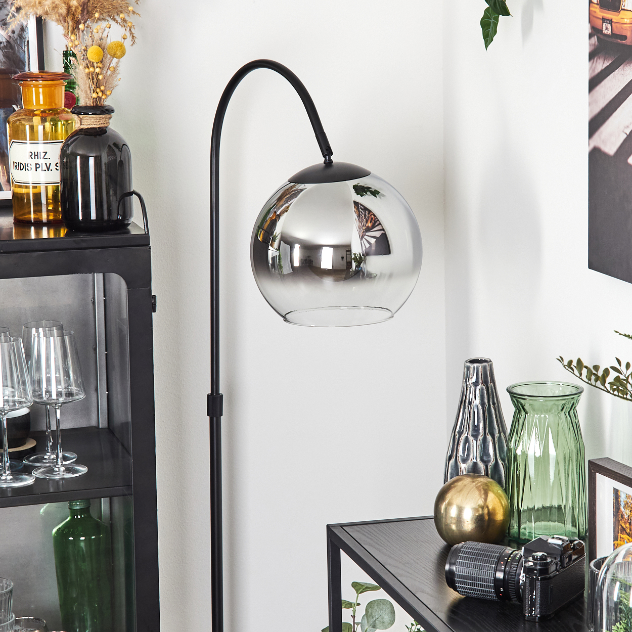 Ein modernes Zimmer mit Stehlampe. Auf einem schwarzen Tisch stehen Vasen, Gläser, eine Kamera und eine goldene Kugel.