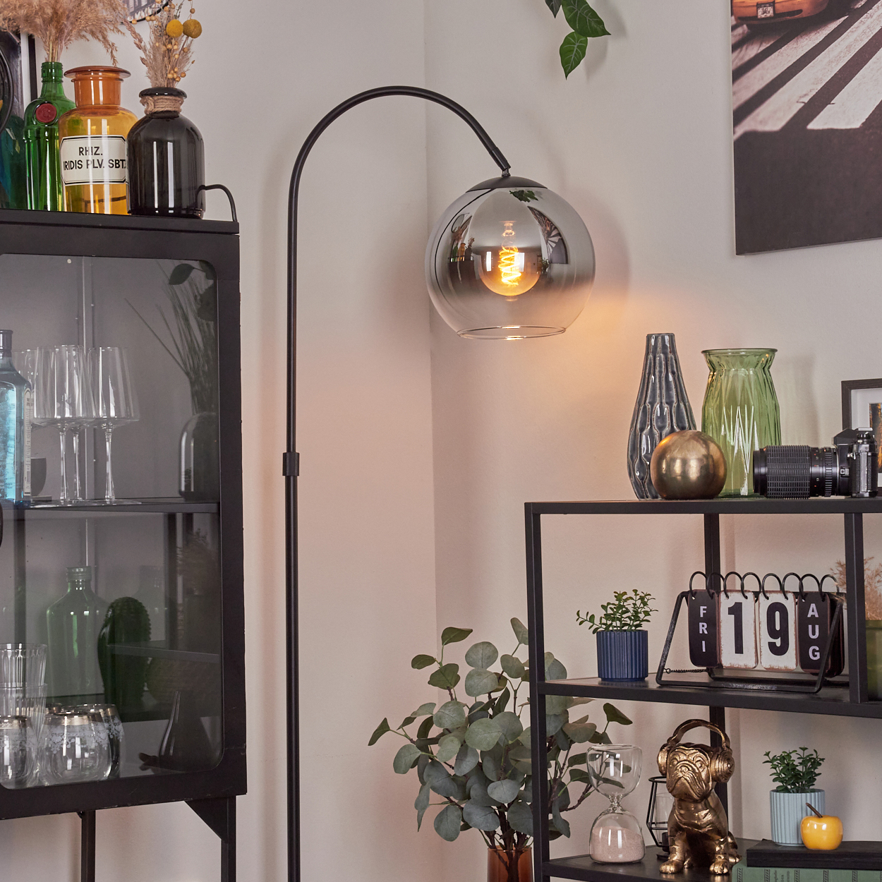 Ein modernes Wohnzimmer mit schwarzer Stehlampe, Glasregalen mit Deko und einem Schrank. Vasen, Pflanzen und ein Kalender sind sichtbar.