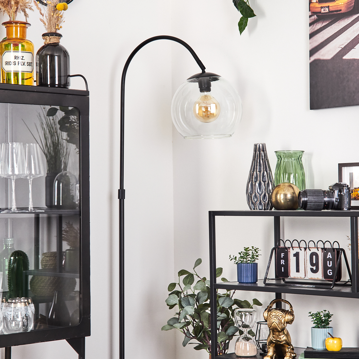 Eine Zimmerecke mit einer schwarzen Stehlampe, Regalen mit Vasen, Pflanzen und einem Kalender. Ein Glasschrank und ein Bild sind sichtbar.