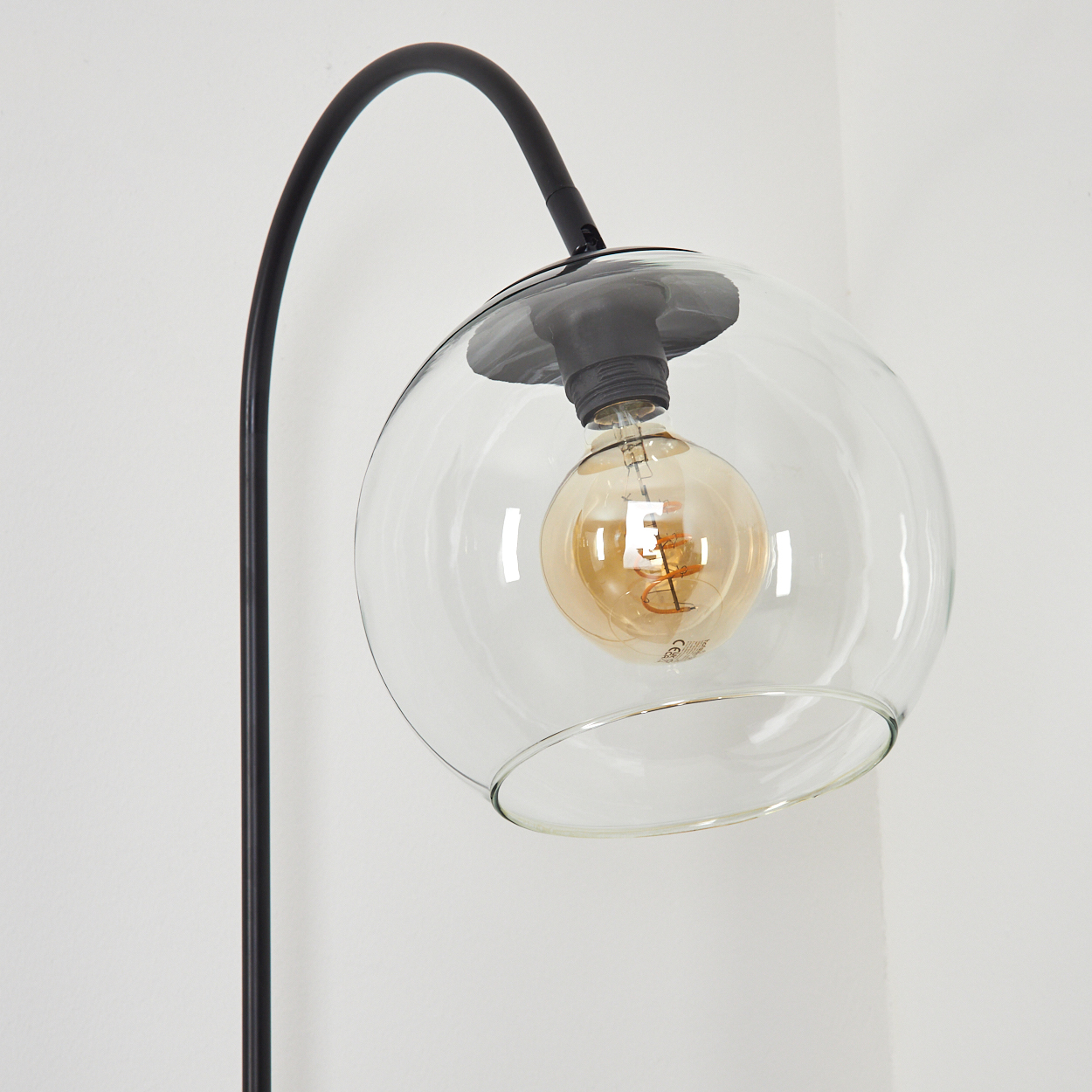 Eine moderne Lampe mit schwarzem, gebogenem Arm, der einen klaren Glaskolben hält. Im Inneren leuchtet eine warmtonige Glühbirne vor einer weißen Wand.