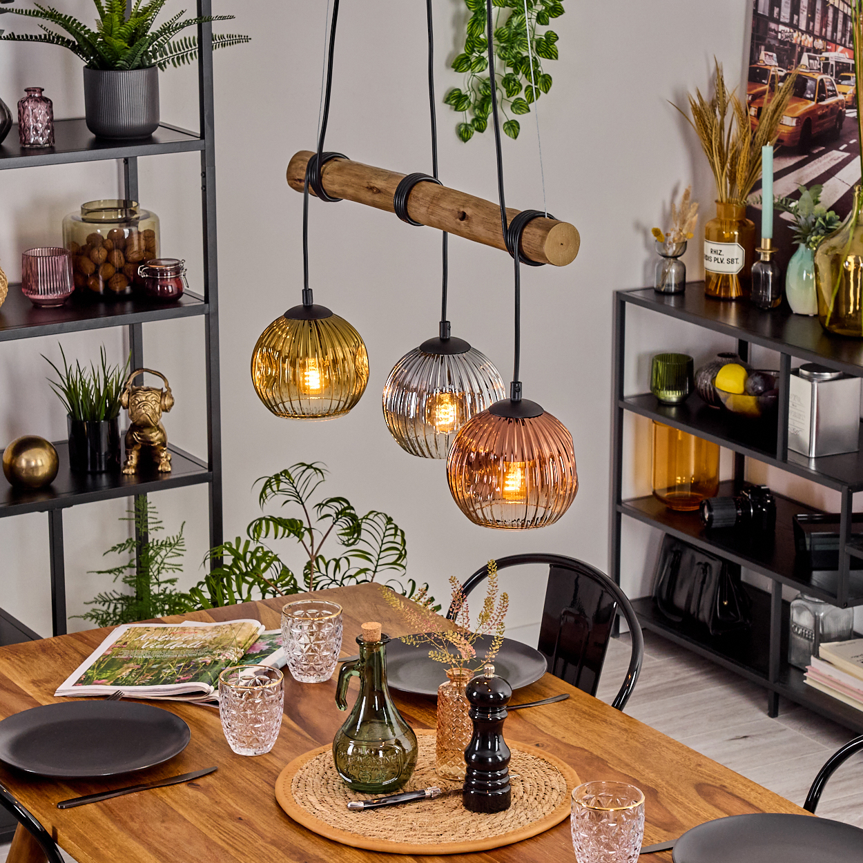 Ein Esszimmer mit Holztisch, drei Pendelleuchten und Regalen. Auf dem Tisch stehen Geschirr und Gläser. Pflanzen und Dekorationen.