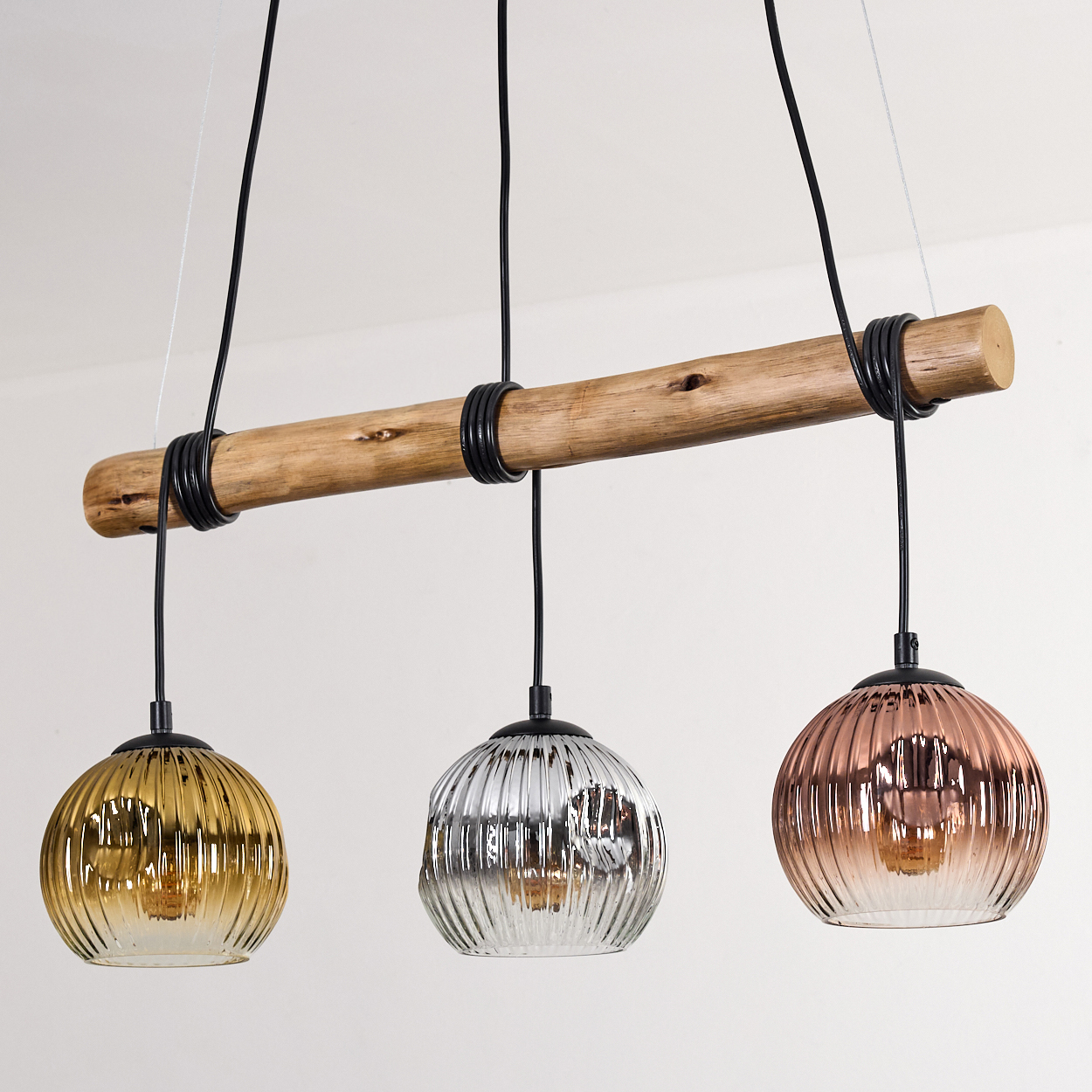 Ein Holzbalken mit drei Pendelleuchten. Jede hat einen unterschiedlich farbigen Glasschirm: Gold, Silber und Roségold.