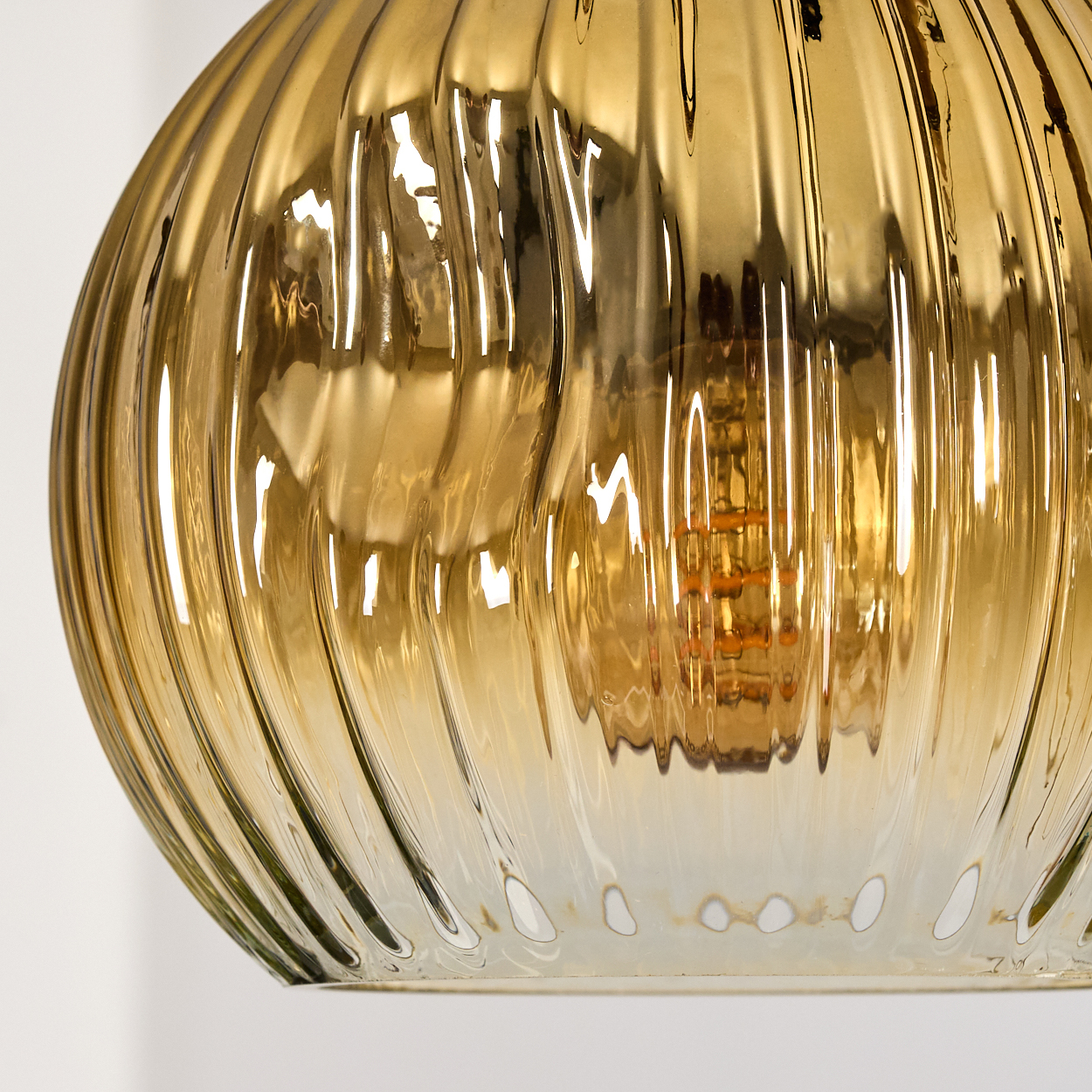 Nahaufnahme eines goldenen Glaslampenschirms mit vertikalen Rippen. Die Glühbirne ist innen sichtbar. Der Hintergrund ist weiß.