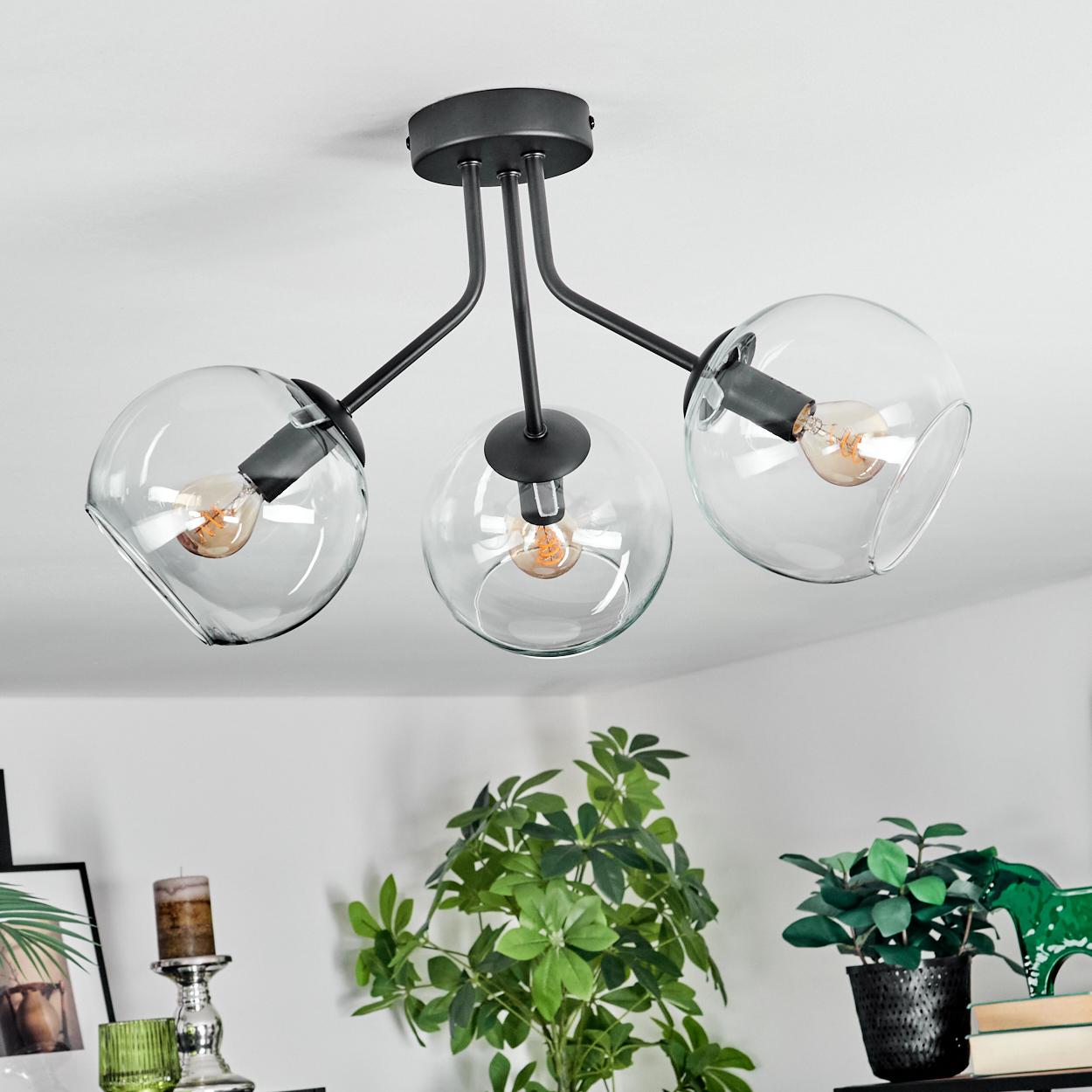 Eine moderne Deckenlampe mit drei Glaskugeln, jede mit einer Glühbirne. Darunter sind Pflanzen und Dekoration zu sehen.