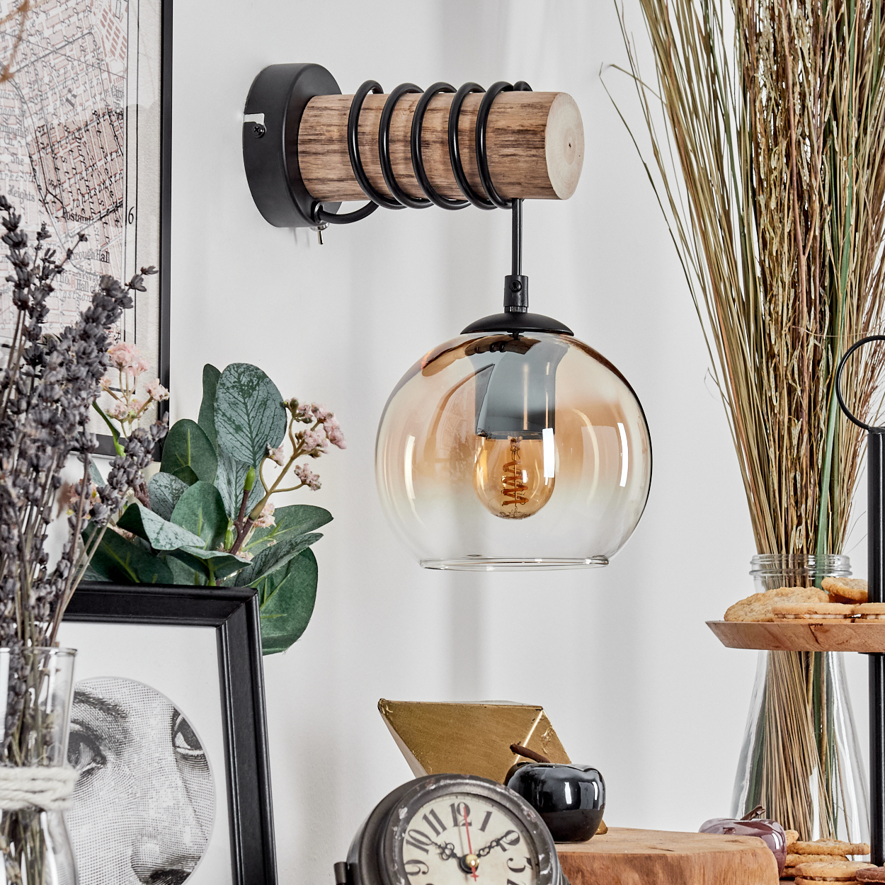 Eine dekorative Wandlampe mit Glasschirm hängt über verschiedenen Dekorationsartikeln. Der Hintergrund umfasst eine Karte und getrocknete Pflanzen.