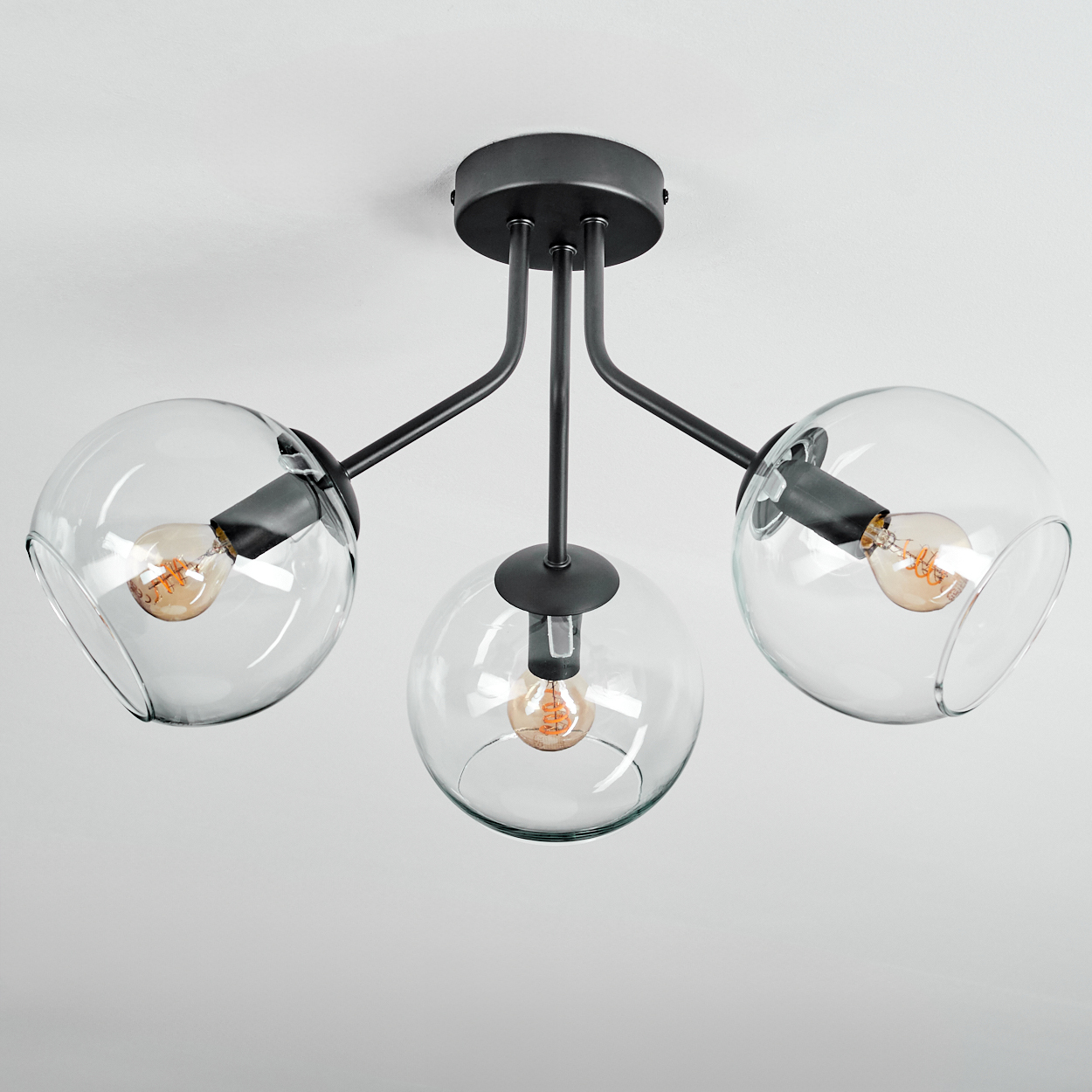 Eine moderne Deckenlampe mit drei Glaskugeln, jede mit einer Glühbirne. Die Halterung ist schwarz und an einer weißen Decke befestigt.