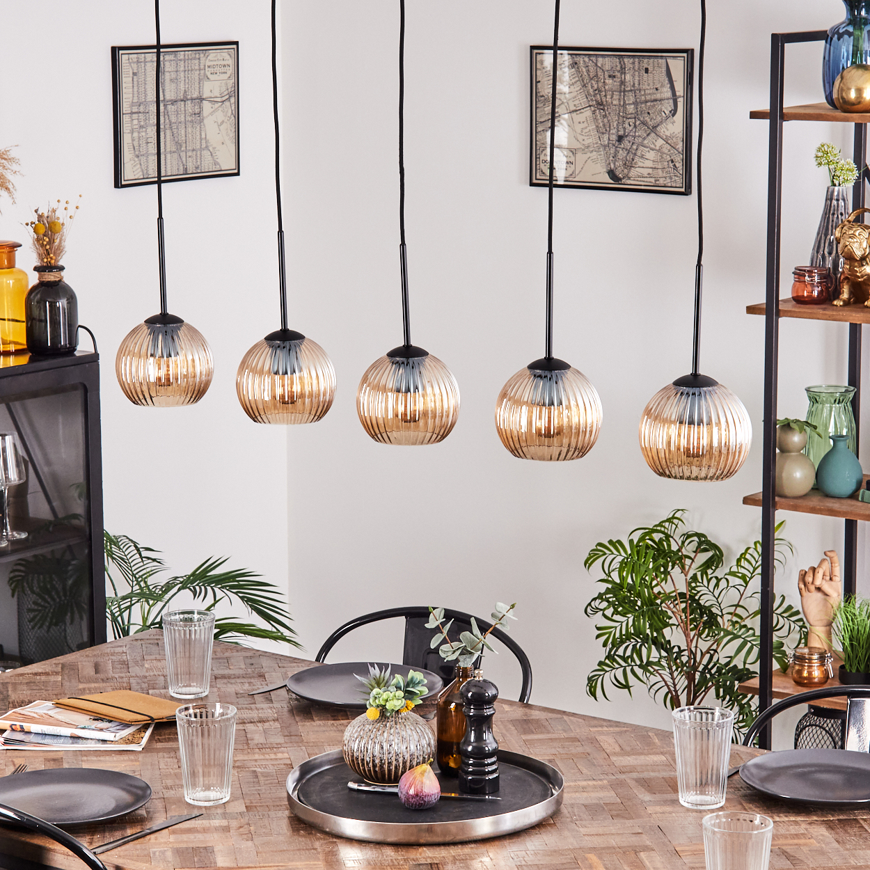 Esszimmer mit Holztisch, runden Lampen, Pflanzen und Dekorationen. Teller, Gläser und ein Tablett mit Deko auf dem Tisch.