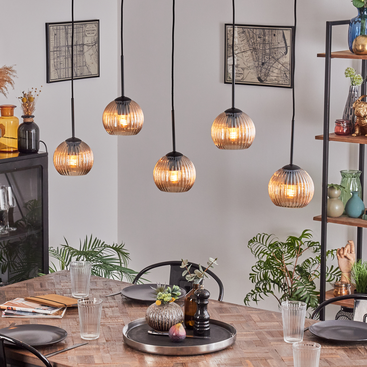 Esszimmer mit Hängelampen. Holztisch mit Tellern, Gläsern und einem Centerpiece. Pflanzen und Regale im Hintergrund.