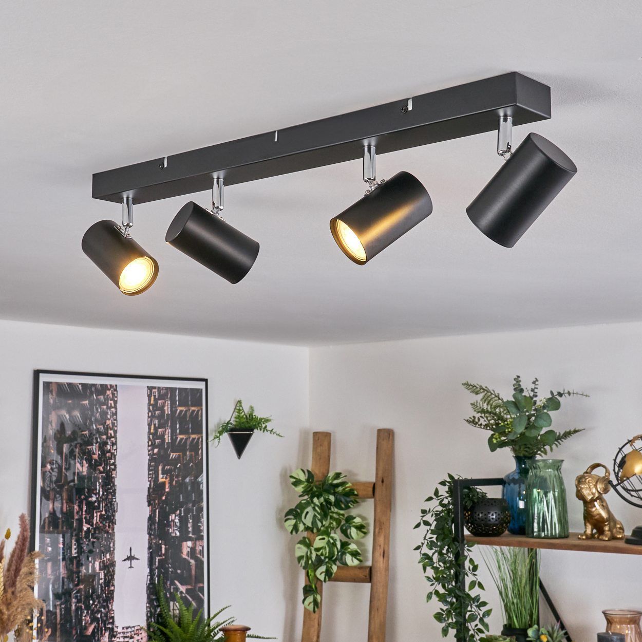 Deckenleuchte mit vier schwarzen Strahlern. Der Raum hat Pflanzen, Kunst und eine Holzleiter. Die Lichter sind an und beleuchten den Raum.