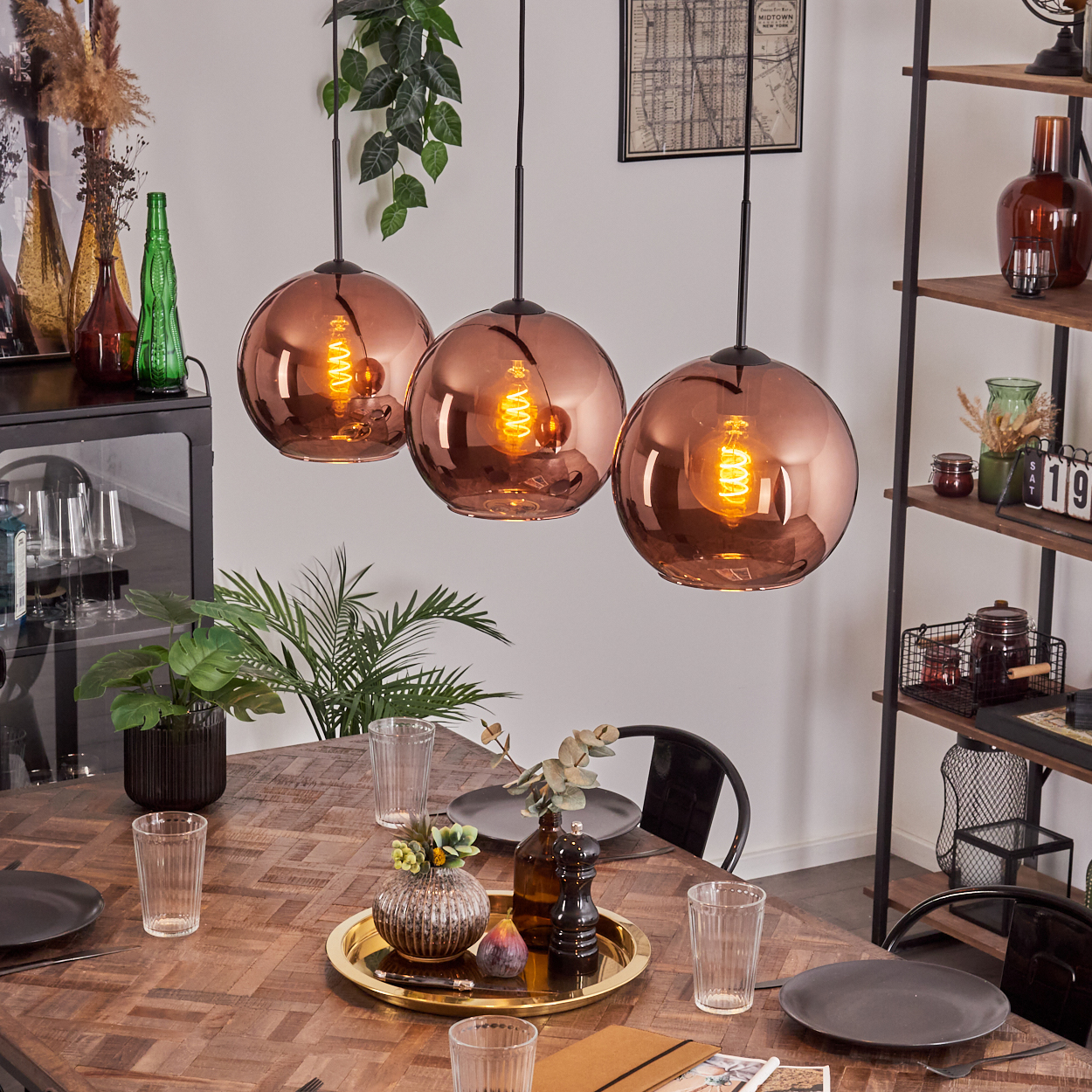 Esszimmer mit Kupfer-Pendelleuchten, Holztisch, Pflanzen und Regalen mit Dekorationen. Teller und Gläser sind gedeckt.
