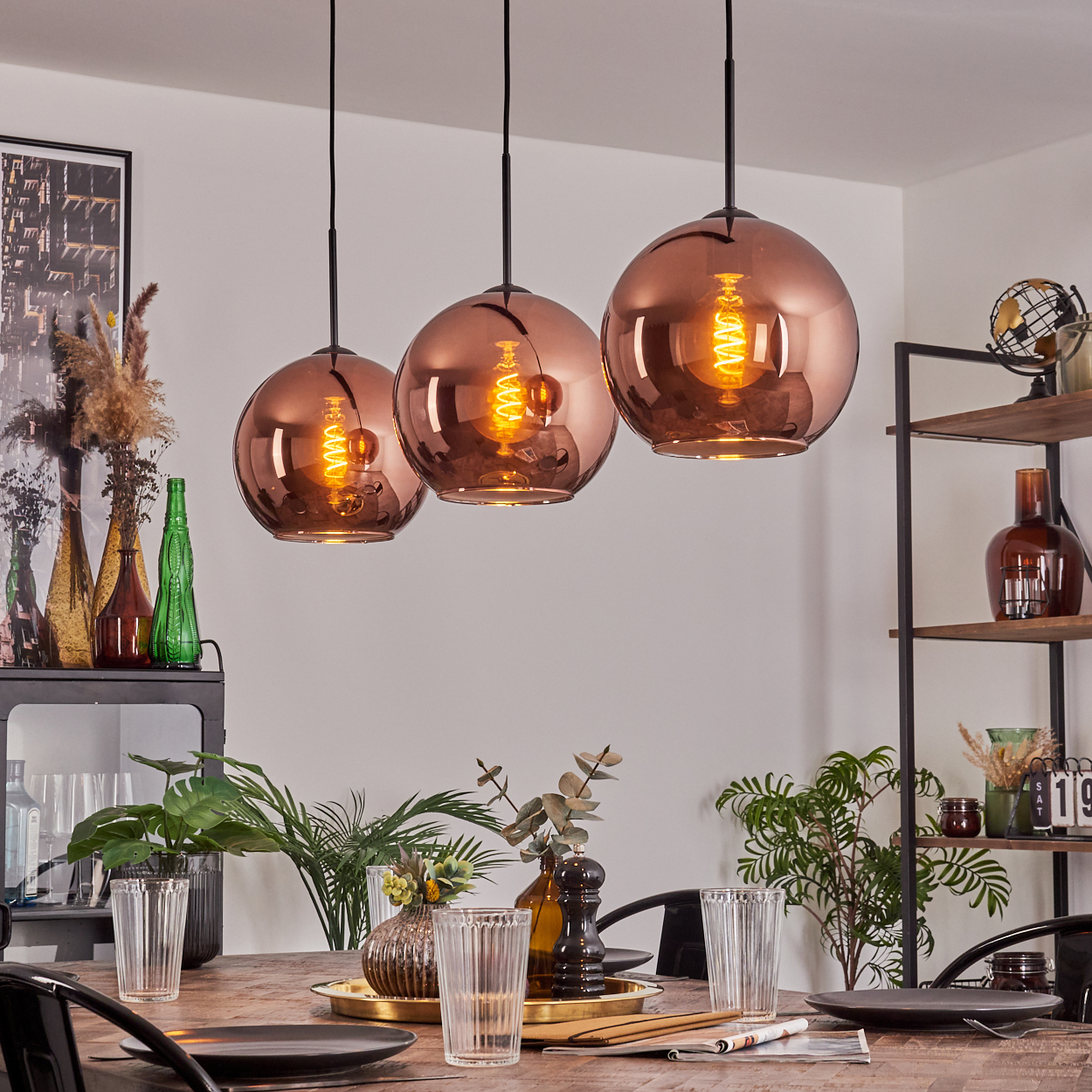 Esszimmer mit Kupfer-Pendelleuchten. Tisch mit Gläsern und Tellern gedeckt. Regale mit Dekoration. Pflanzen und Flaschen sichtbar.