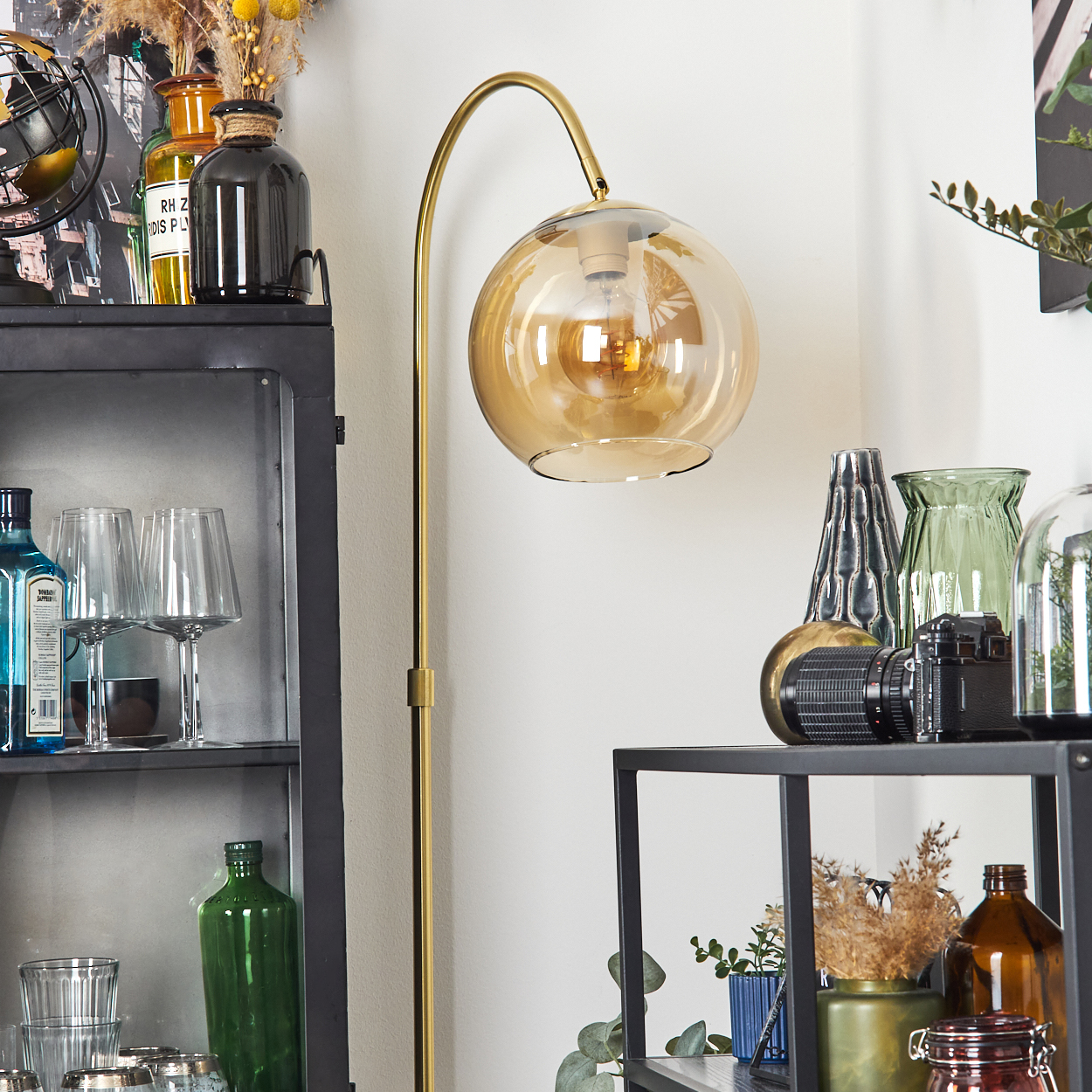 Eine goldene Stehlampe mit kugelförmigem Schirm beleuchtet einen Raum mit dekorativen Flaschen, Gläsern und einer Kamera auf Regalen.