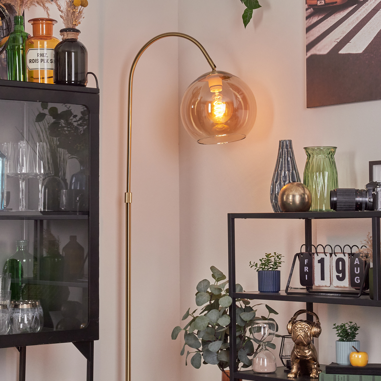 Eine goldene Stehlampe mit kugelförmigem Schirm beleuchtet einen Raum mit Dekorregalen. Eine Bulldoggenstatue trägt Kopfhörer.