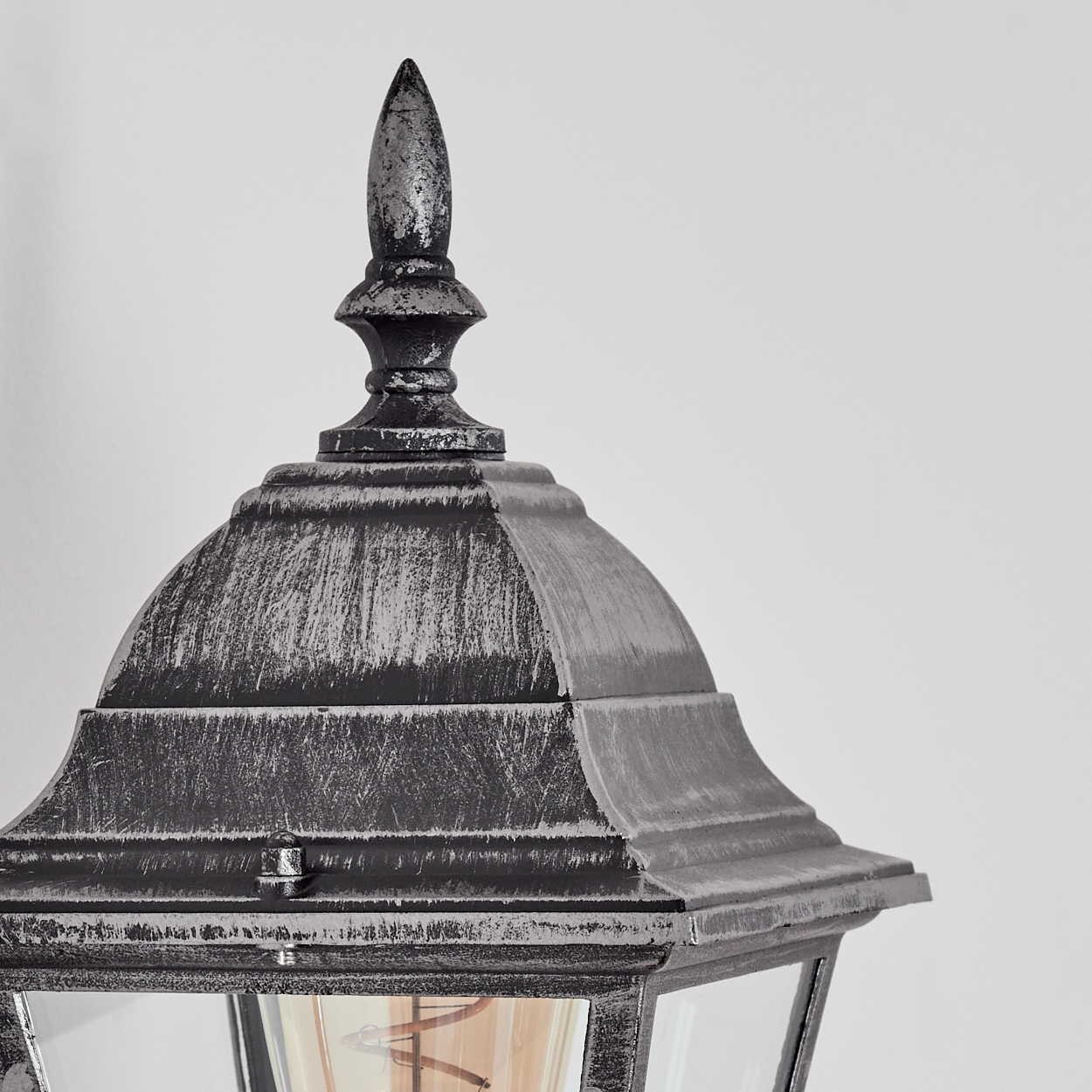 Nahaufnahme einer verzierten, schwarz-silbernen Außenlampe. Die Lampe hat eine spitze Oberseite und ein Glasgehäuse mit einer sichtbaren Glühbirne.