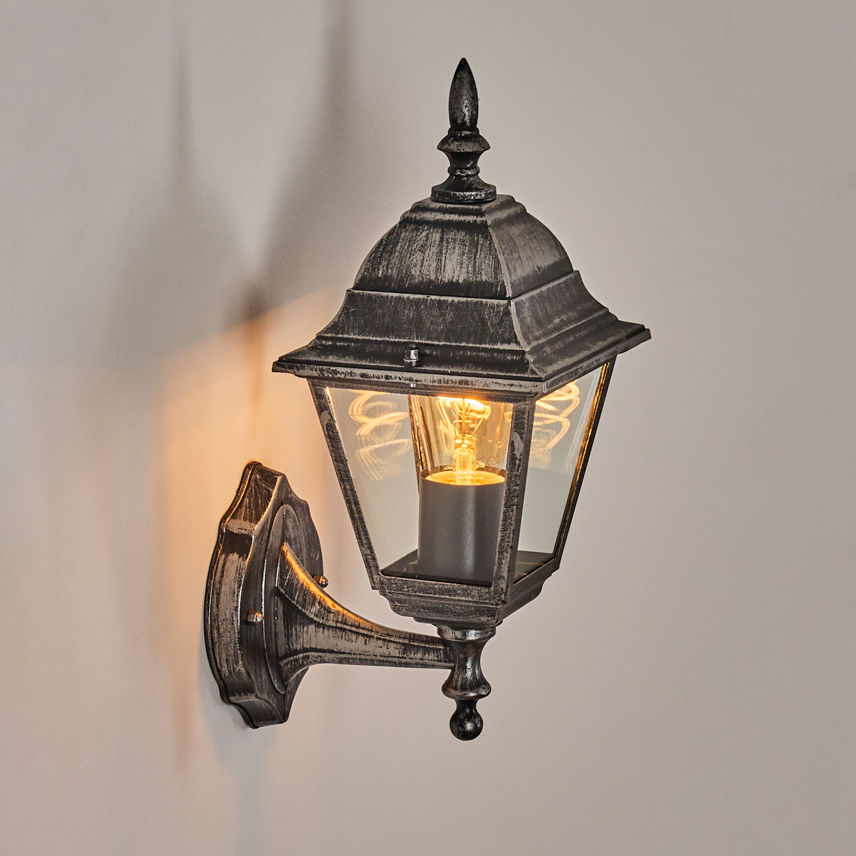 Eine verzierte, silberne Wandlampe mit einer leuchtenden Glühbirne. Sie ist an einer hellen Wand montiert.