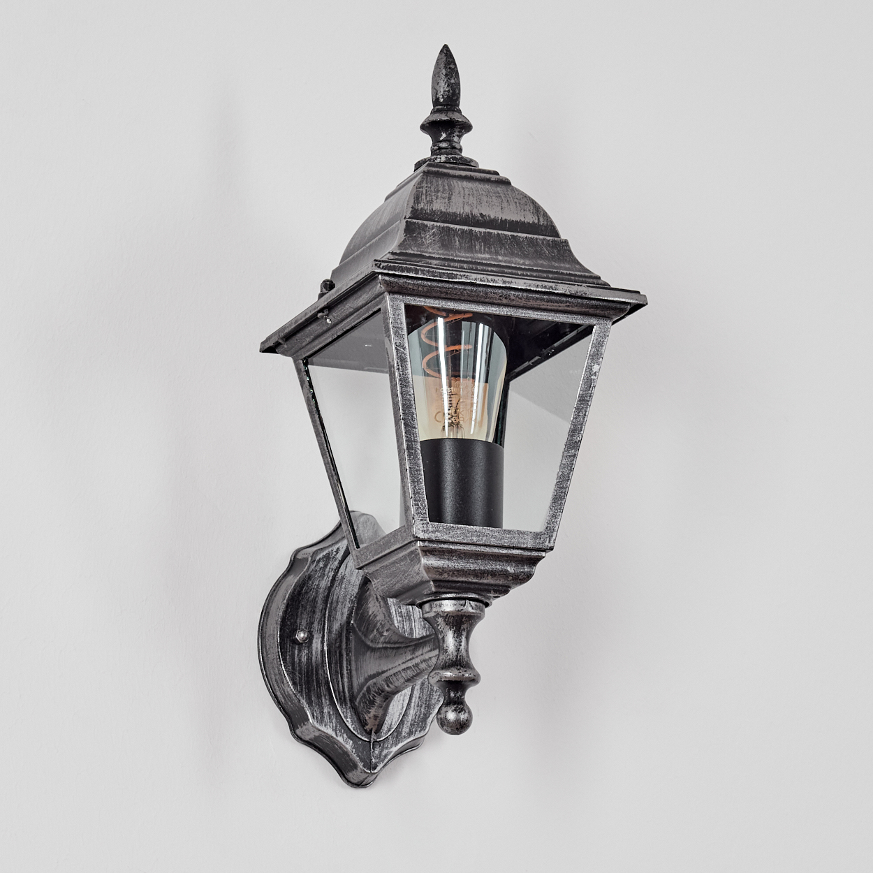 Eine dekorative Außenwandleuchte mit silbernem Finish. Sie hat ein Glasgehäuse und eine dekorative Glühbirne. An einer weißen Wand montiert.
