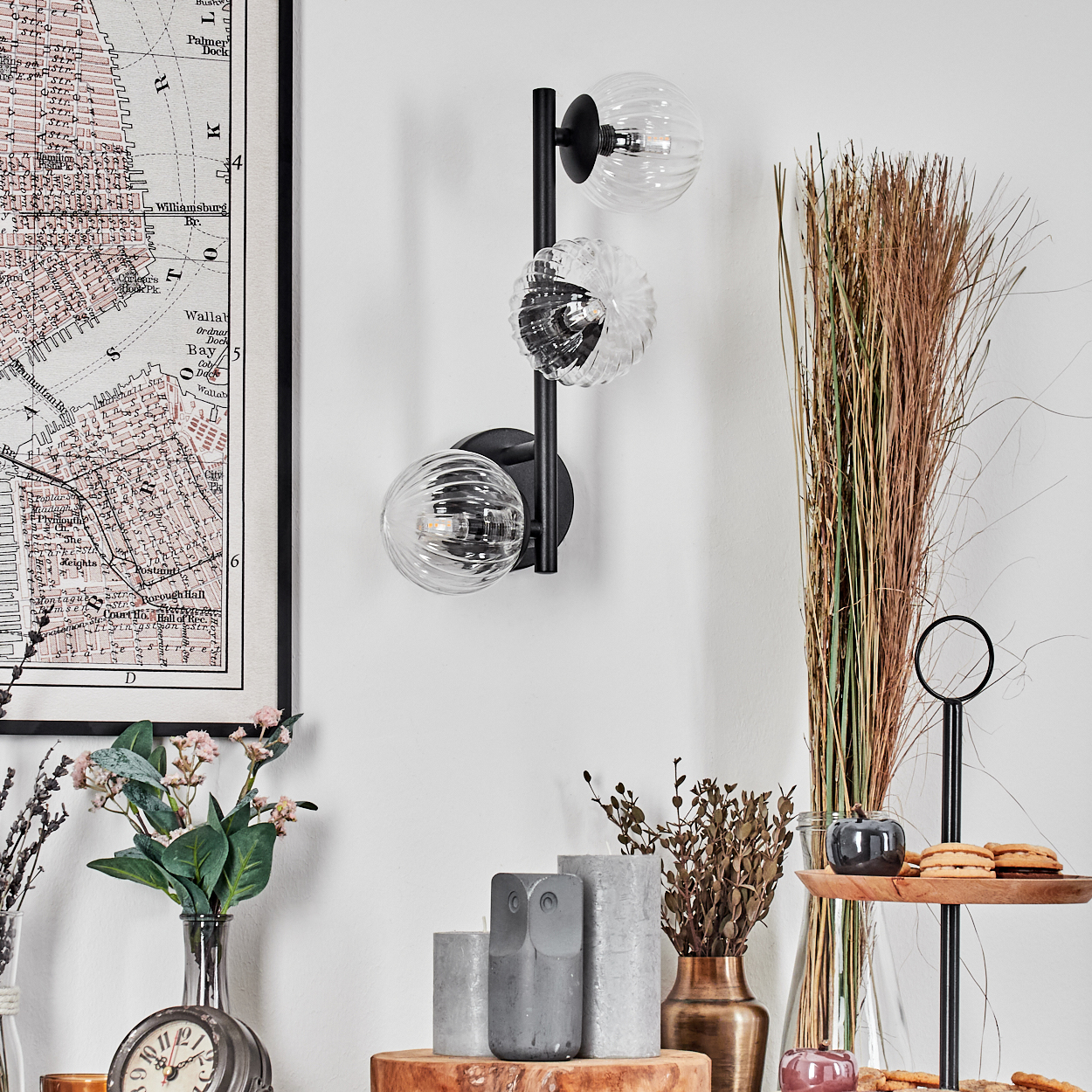 Eine Wandlampe mit drei Glaskugeln ist an einer weißen Wand montiert. Darunter befinden sich Dekorationen, darunter eine Karte, Blumen und Kerzen.
