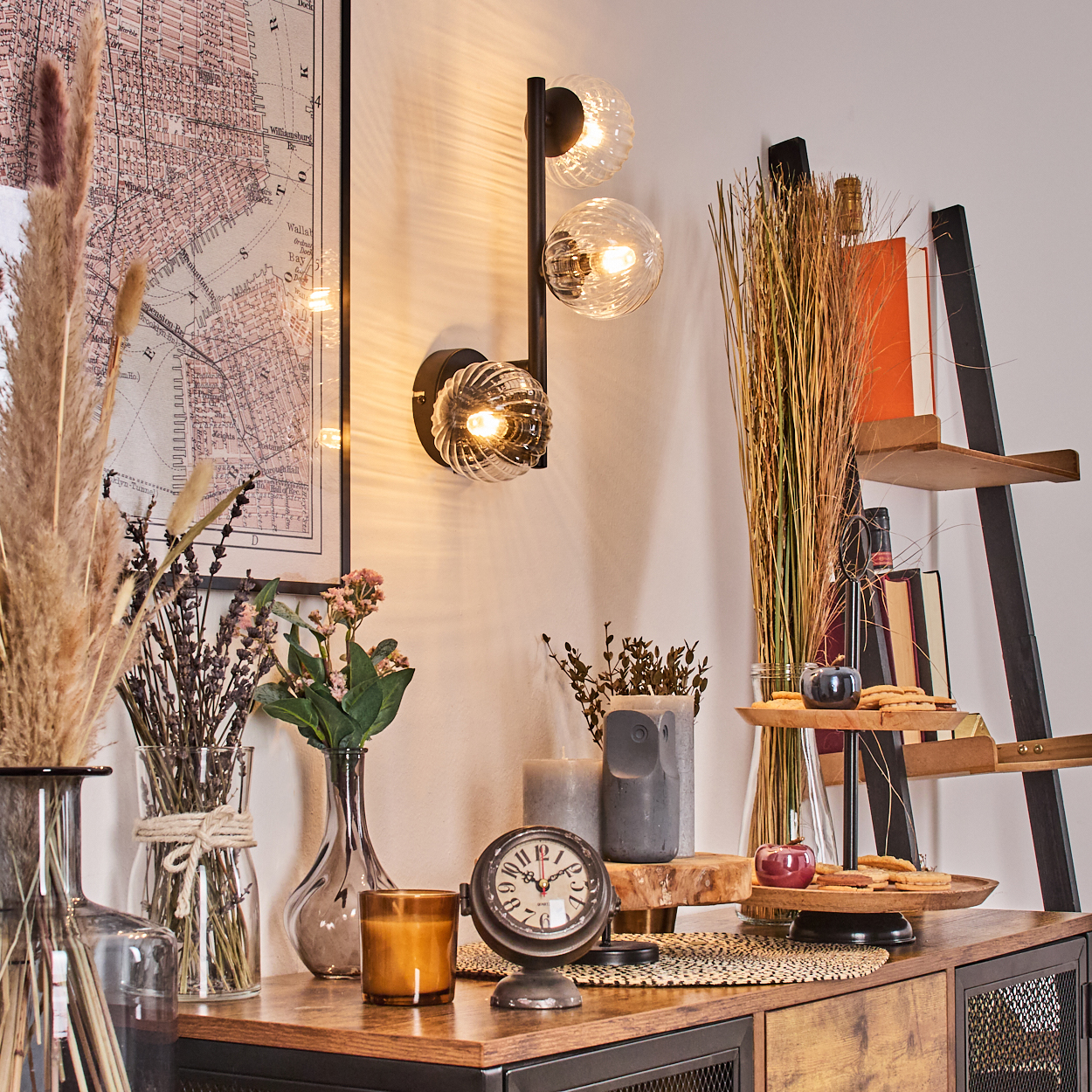 Ein Raum mit Wandlampe, Vasen mit Blumen, einer Uhr und einem Schrank mit Dekorationsartikeln. An der Wand hängt eine Karte.