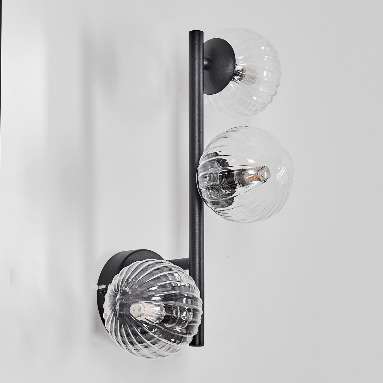 Eine moderne Wandlampe mit drei Glaskugelschirmen. Die Halterung ist schwarz, mit klaren, geriffelten Glasschirmen. An einer weißen Wand montiert.