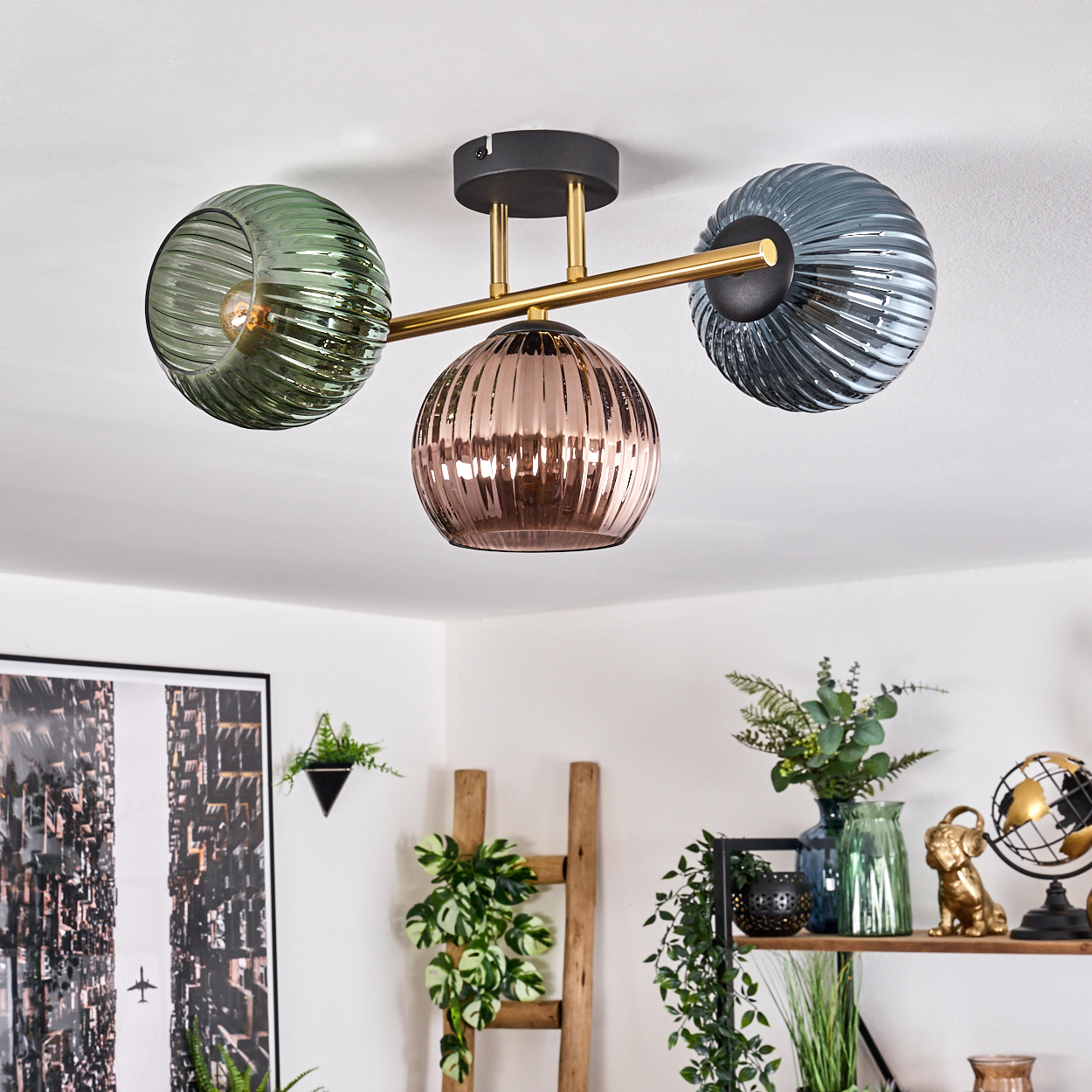 Eine moderne Deckenleuchte mit drei Glasschirmen in Grün, Rosa und Blau. Deko umfasst Pflanzen, eine Leiter und einen Globus.