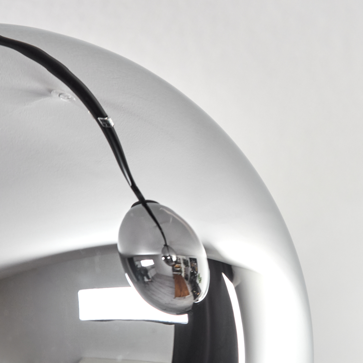 Eine verchromte, reflektierende Lampe mit schwarzem Kabel hängt vor weißem Hintergrund. Die Reflexion zeigt einen Raum mit Möbeln.
