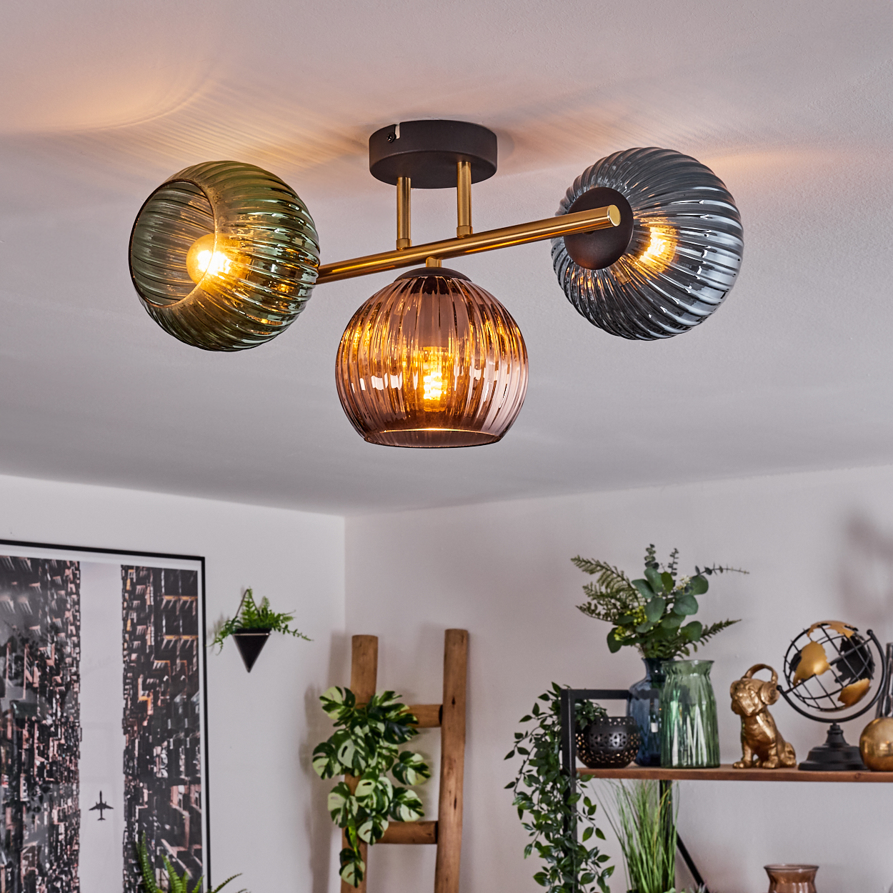 Eine moderne Deckenleuchte mit drei Glaskugeln. Darunter ein Regal mit Dekoration und Pflanzen. Ein großes Bild und eine Leiter sind ebenfalls sichtbar.