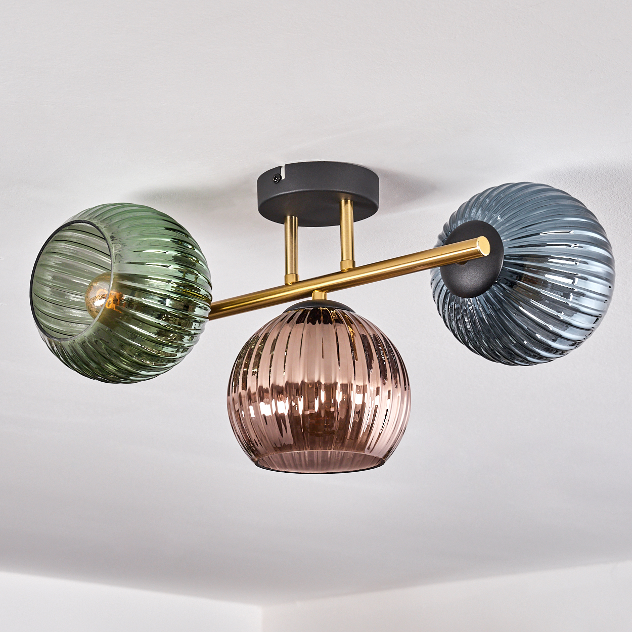 Eine moderne Deckenleuchte mit drei Glaskugeln in Grün, Rosa und Blau. Goldene Arme, schwarze Basis und weiße Decke.