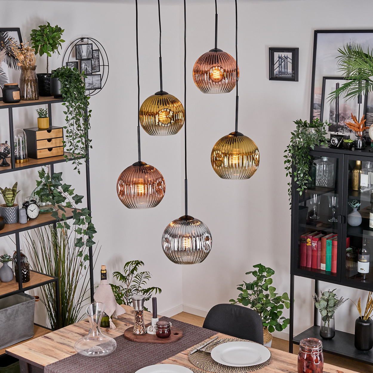 Esszimmer mit Hängekugelleuchten. Tisch mit Tellern, Karaffe und Kerzen. Regale mit Pflanzen, Dekoration und einem schwarzen Schrank.
