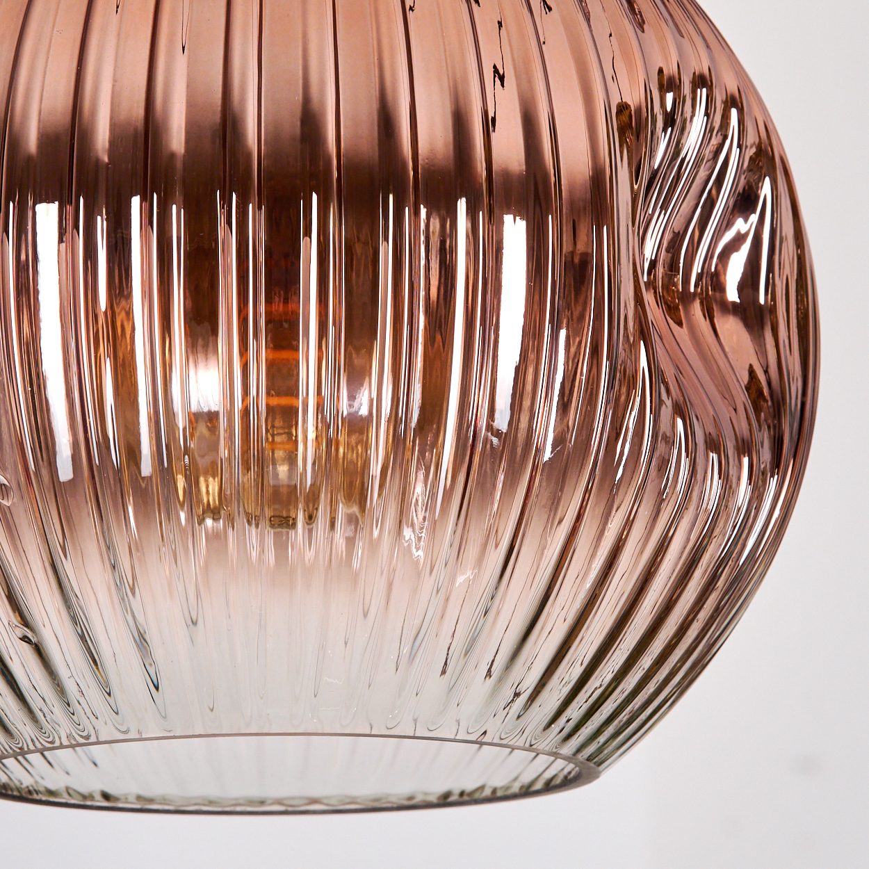 Nahaufnahme eines dekorativen Lampenschirms aus Glas. Er ist geriffelt und hat einen roségoldenen Farbton, der das Licht reflektiert. Der Hintergrund ist weiß.
