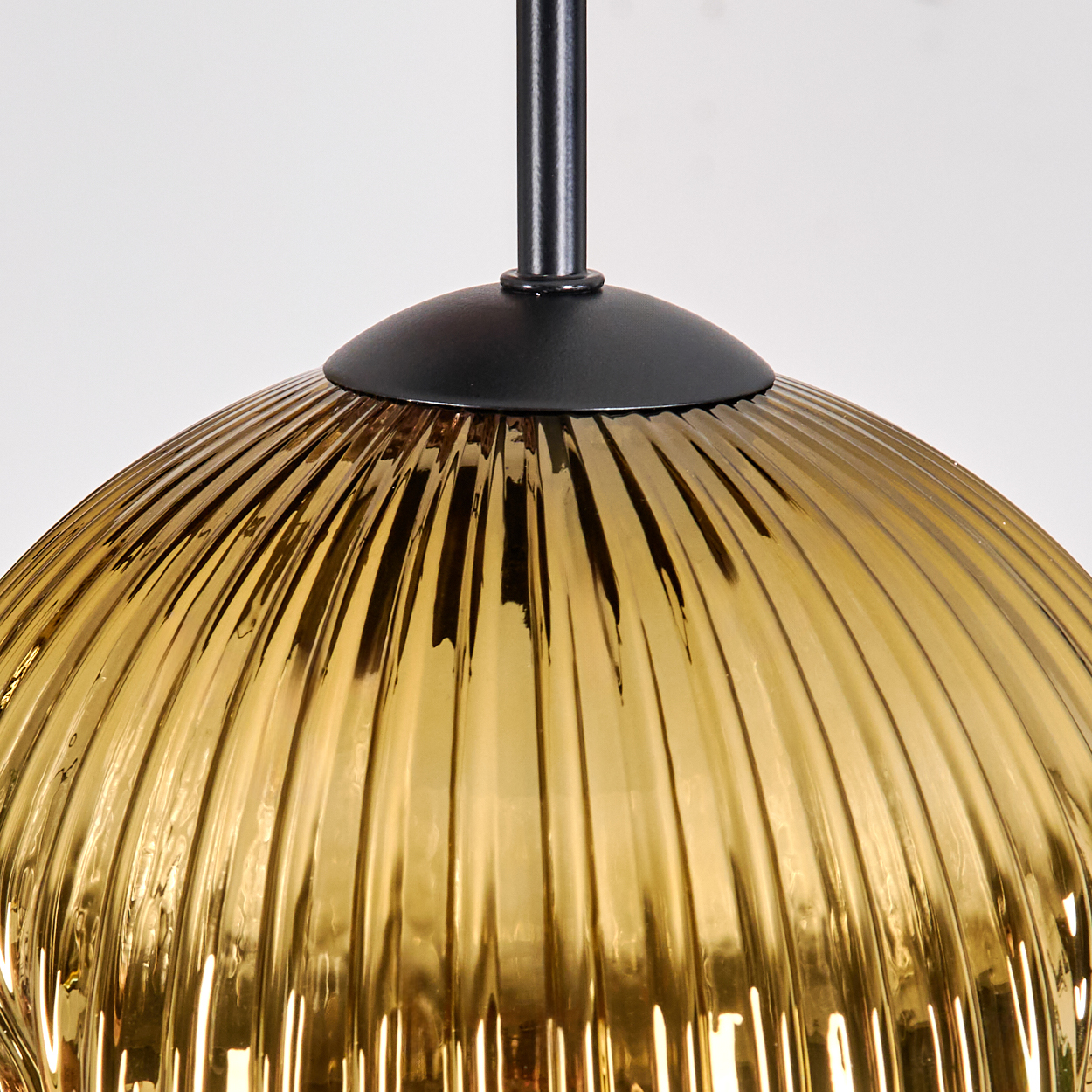Nahaufnahme eines goldenen Glaslampenschirms mit vertikalen Rippen, der an einer schwarzen Halterung vor weißem Hintergrund hängt.