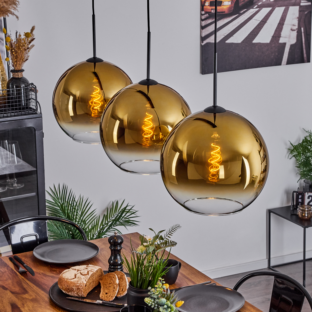 Drei goldene Hängelampen über einem Esstisch mit Tellern, Brot und Pflanzen. Ein Bild hängt an der Wand.