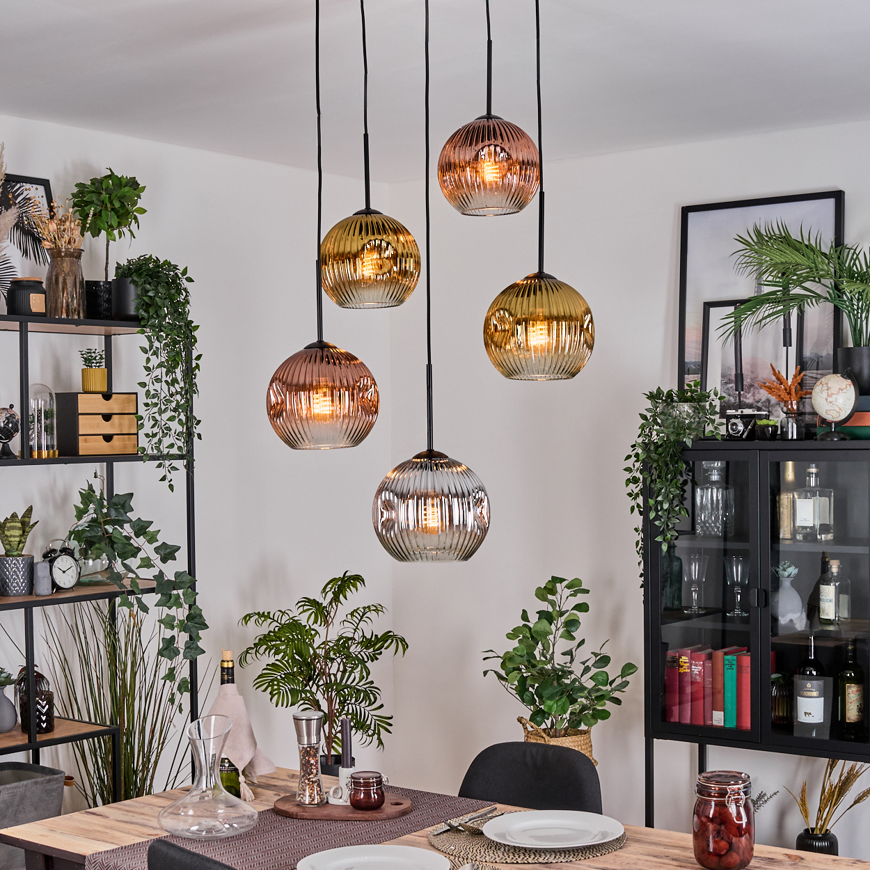 Esszimmer mit Holztisch, Hängelampen, Pflanzen und einem schwarzen Schrank. Die Lampen haben Glasschirme in verschiedenen Farben.