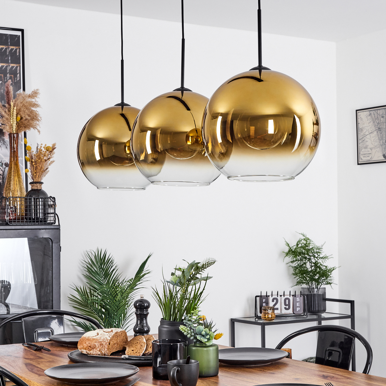Esszimmer mit drei goldenen Pendelleuchten, einem Holztisch mit Tellern und Pflanzen.
