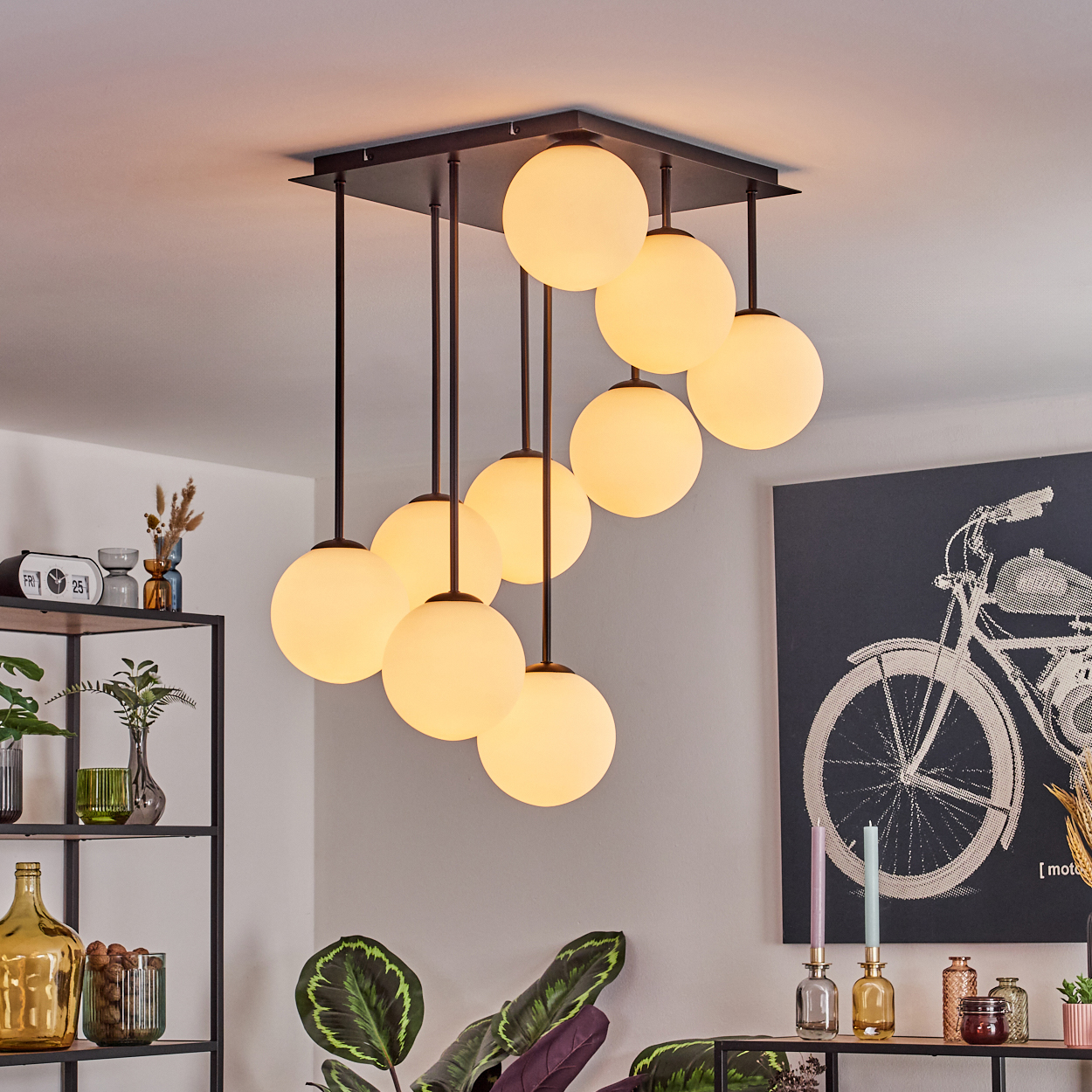 Moderne Deckenleuchte mit neun kugelförmigen Glühbirnen. Pflanzen und ein Regal mit Deko im Vordergrund. Ein Gemälde an der Wand.