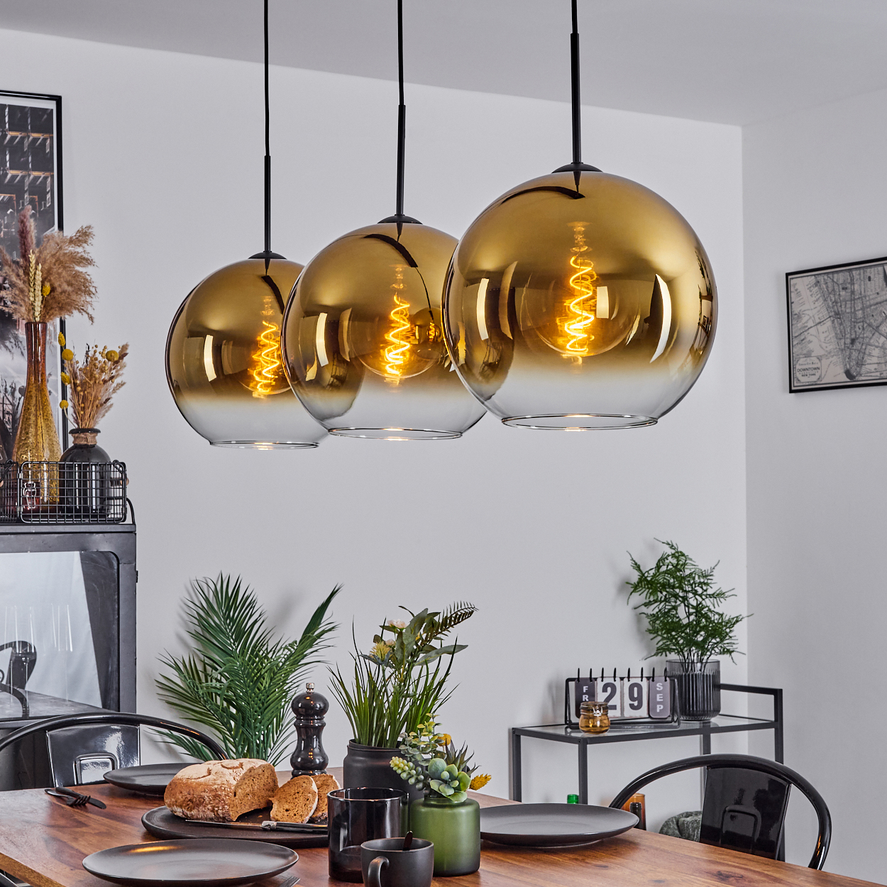Drei goldene Hängelampen über einem Holztisch, der für eine Mahlzeit gedeckt ist. Brot, Pflanzen und schwarze Teller sind auf dem Tisch.