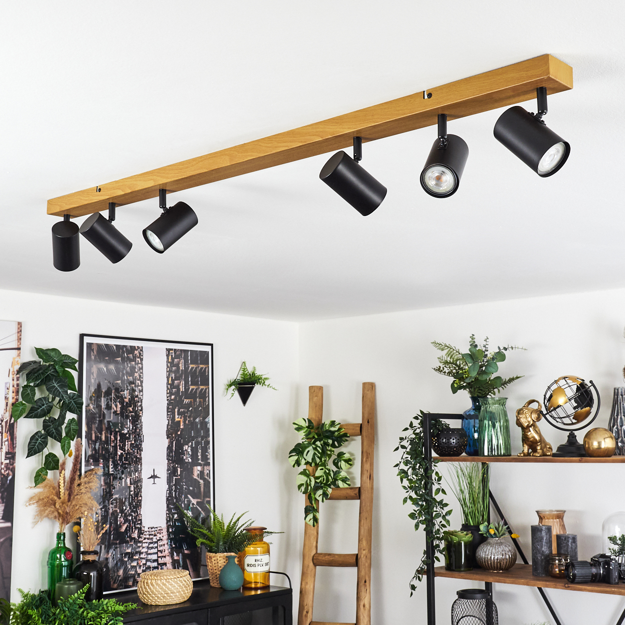 Ein modernes Zimmer hat eine Deckenleuchte aus Holz mit schwarzen Strahlern. Darunter sind Pflanzen, eine Leiter und Regale mit Dekorationen.